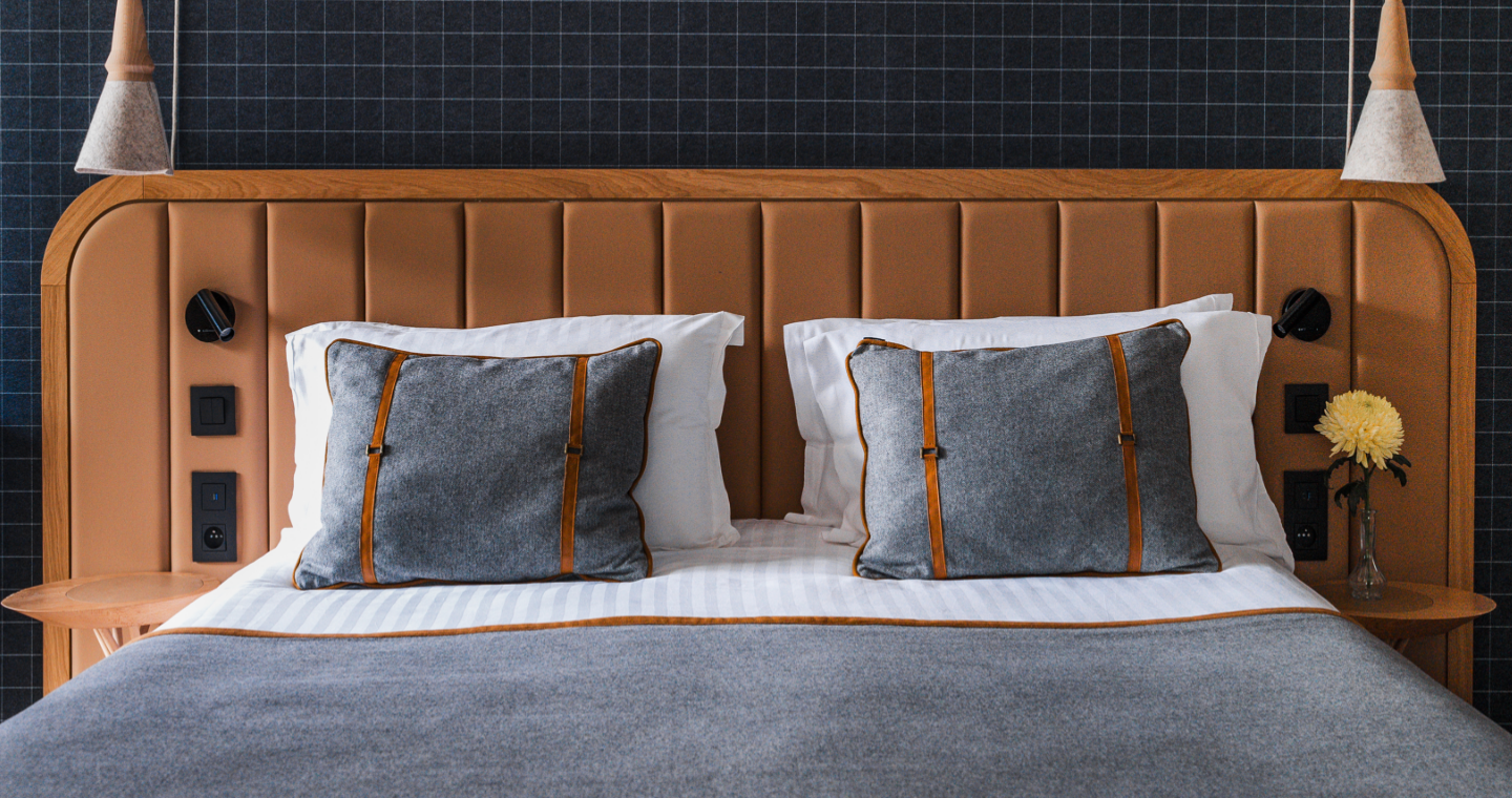 Double bed with brown leather headboard, two gray cushions with leather straps, and a gray blanket.