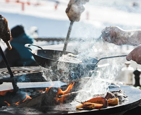 Le brasero du restaurant Le M avec différentes viandes et légumes cuisant à la chaleur des flammes, tandis qu'un cuisinier les arrose.