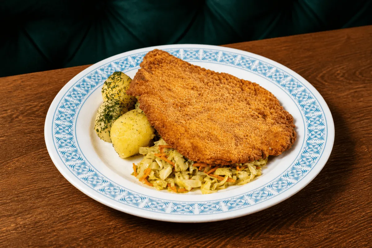 Duży, klasyczny kotlet schabowy w złocistej panierce, podany na białym talerzu z niebieskim wzorem. Obok mięsa leżą ugotowane ziemniaki posypane koperkiem oraz porcja zasmażanej kapusty z marchewką. Danie stoi na drewnianym stole w restauracji, w tle widać ciemnozielone oparcie kanapy.