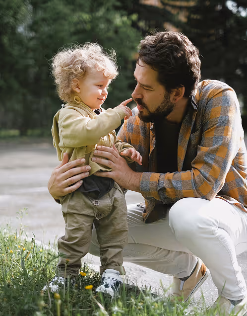 Man crouching outdoors smiling as toddler with curly hair touches his nose.