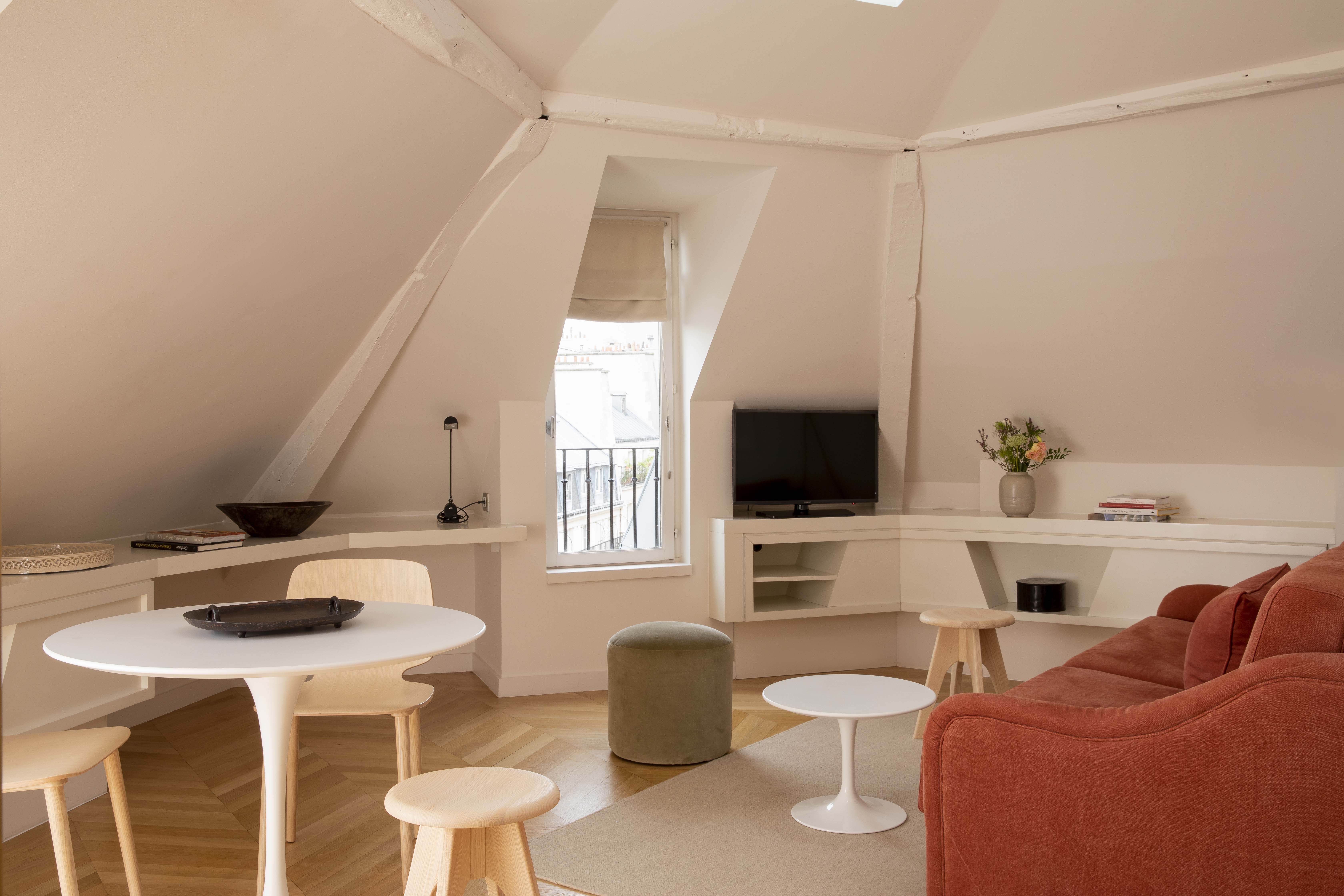 Attic living room with a red sofa, a white coffee table, a green pouf, a small round table with light wood chairs, and a television in front of a window.