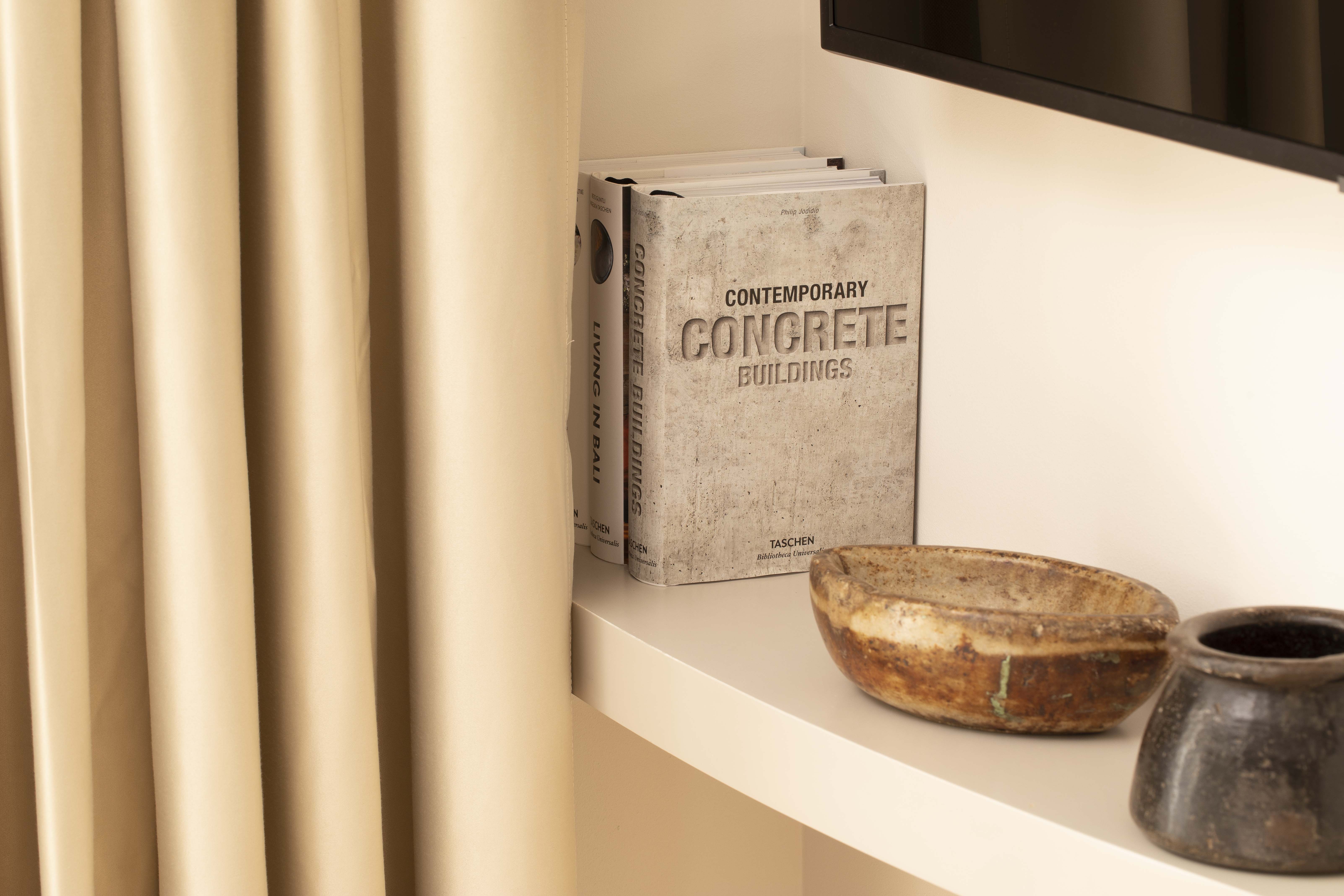 White shelf with books on architecture and two decorative ceramic bowls, next to beige curtains.