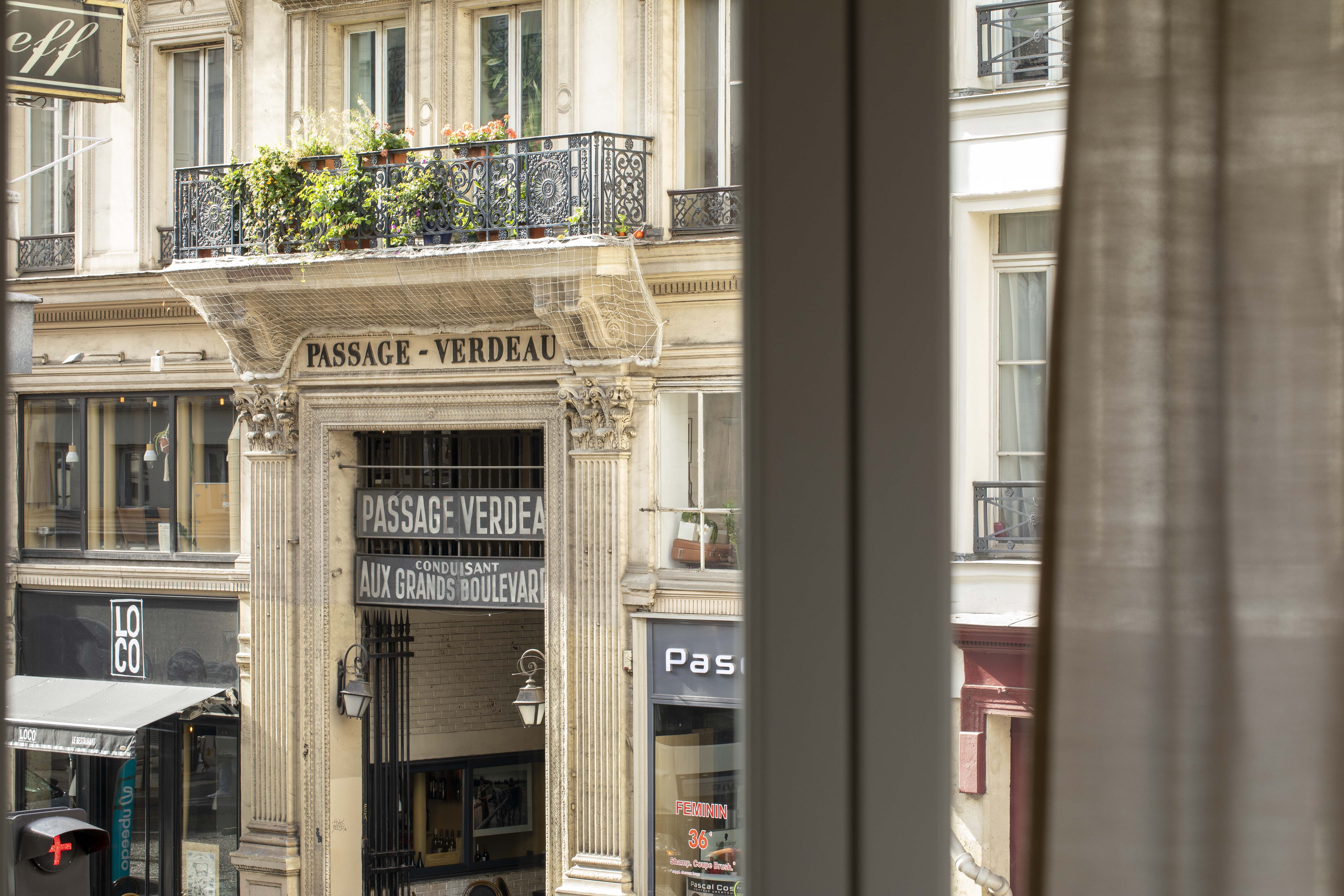 Entrance to Passage Verdeau in Paris with a stone facade and a flowered balcony, partial view through a window with a curtain.