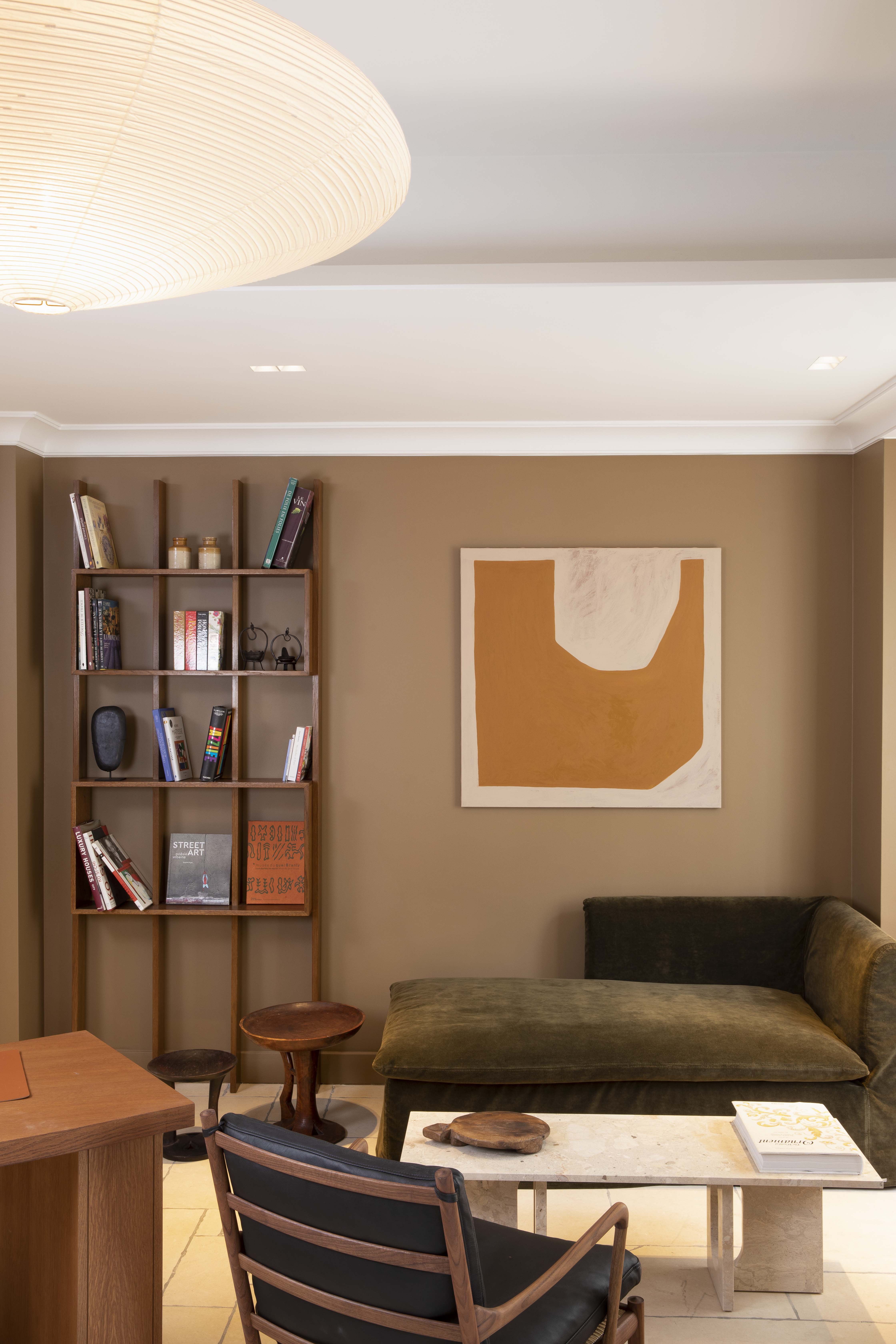 Modern living room with a green sofa, a wooden chair with a black cushion, a stone coffee table, a wall shelf with books, and an orange abstract painting on a beige wall.