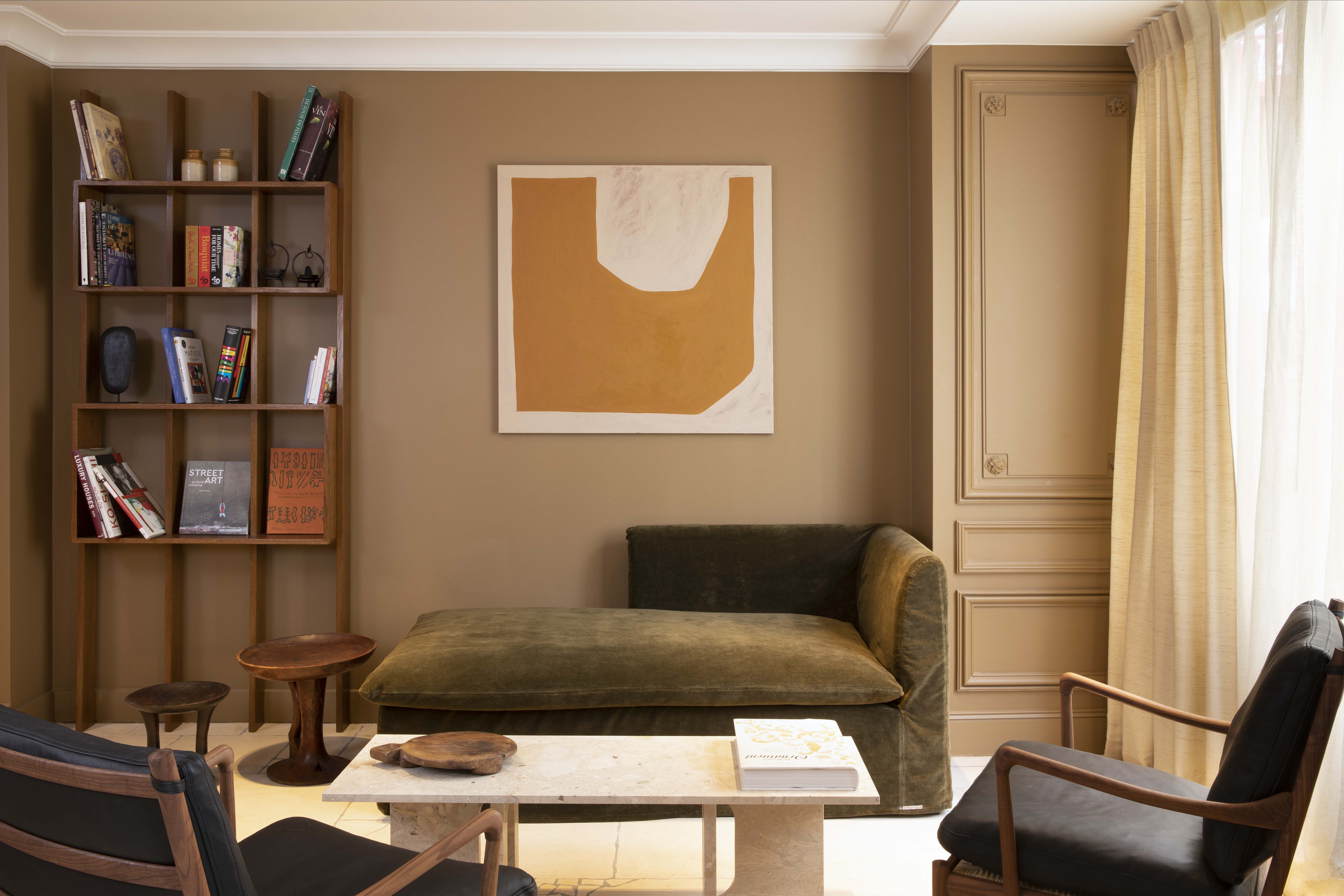Salon avec canapé vert olive, deux fauteuils en bois et cuir noirs, table basse en pierre, étagère en bois avec livres, et tableau abstrait orange sur mur beige.