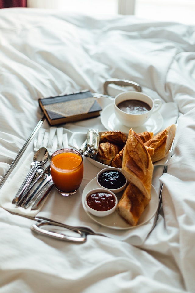 Breakfast tray on a white bed with orange juice, coffee, croissants, baguette, and jams.