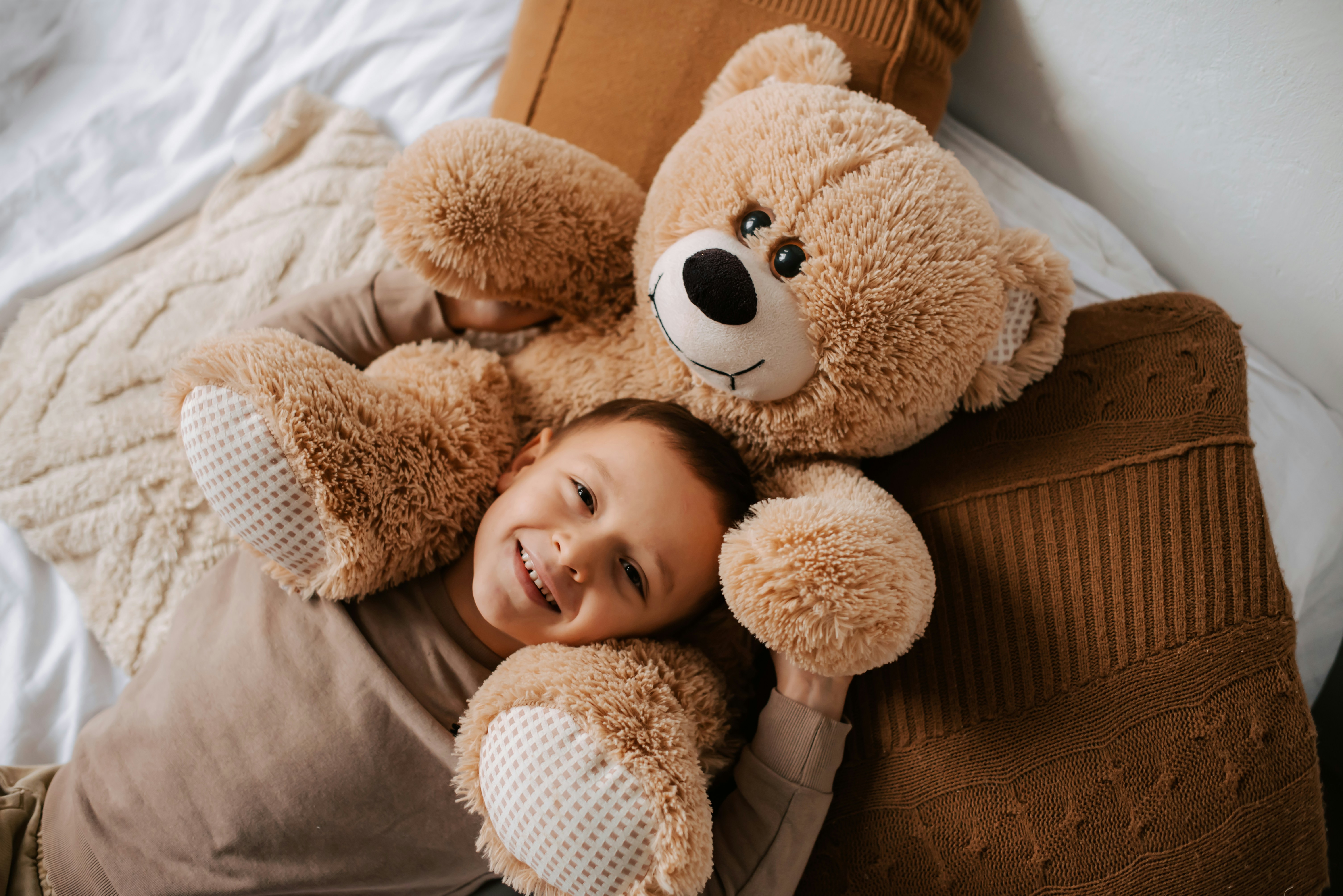 A smiling boy lying on a bed cuddling a large brown teddy bear.