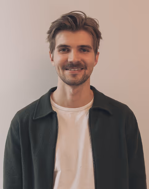 Ung mann med kort brunt hår og skjegg smiler, iført hvit t-skjorte og mørk jakke foran en lys vegg.