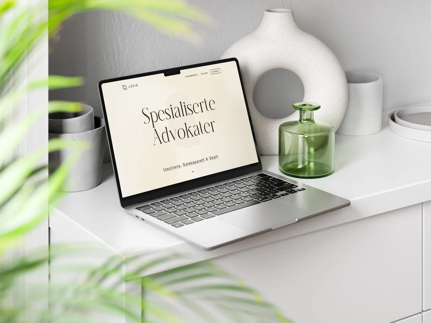Bærbar datamaskin på hvit kommode med nettside for spesialiserte advokater vist på skjermen, omgitt av grønne blad og dekorative keramiske og glassvaser.