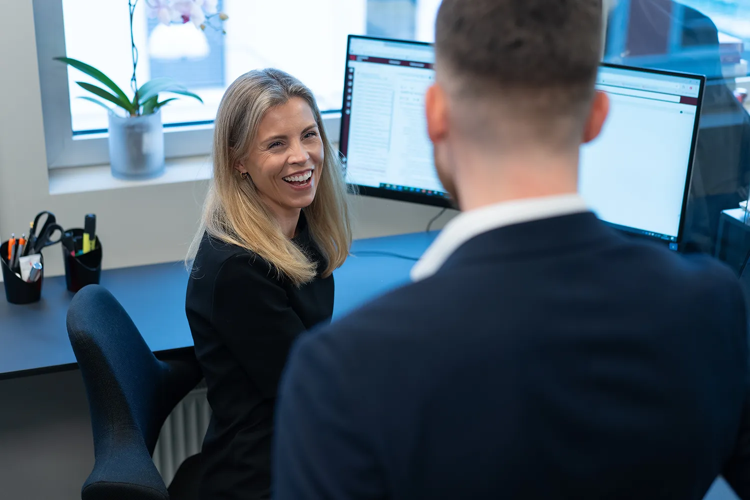 Kvinne smiler og snakker med en mann i en kontorsetting med dataskjermer i bakgrunnen.