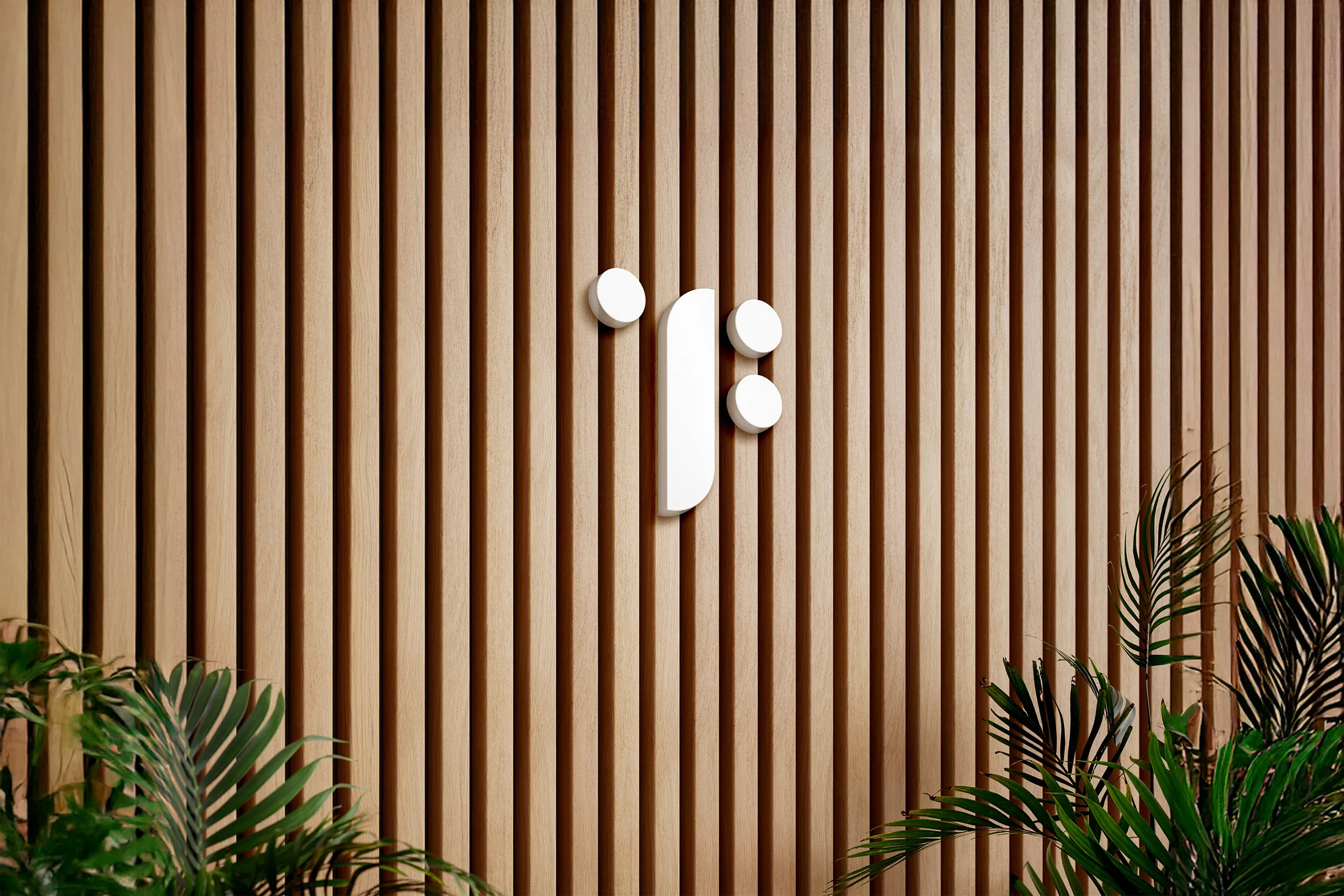 Hvit minimalistisk logo på en vertikal trepanelvegg med grønne planter i forgrunnen.