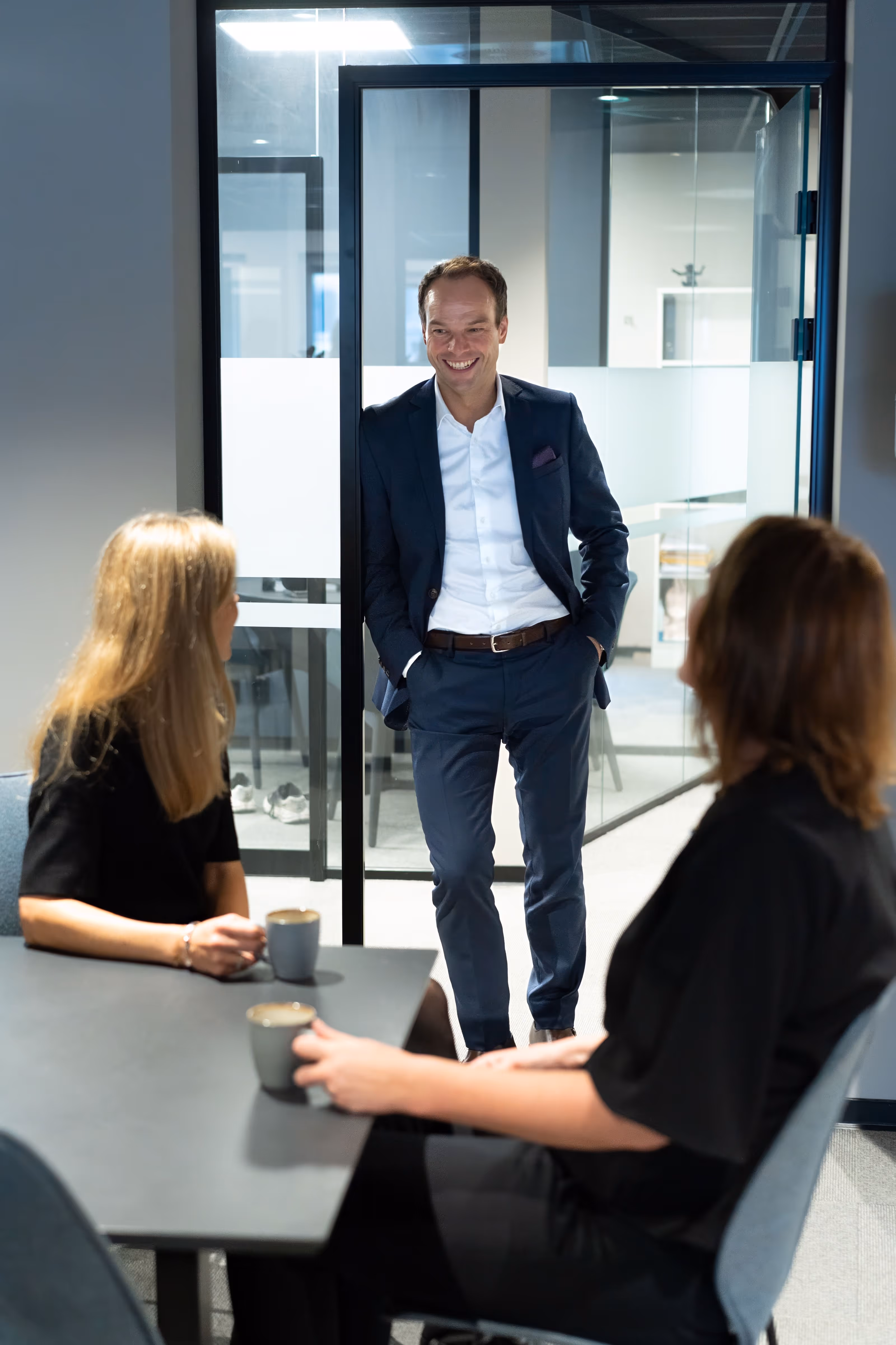 En mann i dress smiler og snakker med to kvinner som sitter ved et bord med kaffekopper i et kontormiljø.
