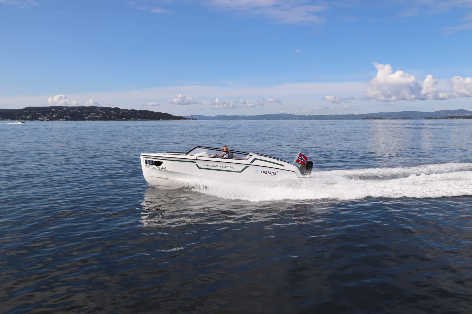 Elektrisk motorbåt med norsk flagg som kjører på sjøen ved kystlandskap.