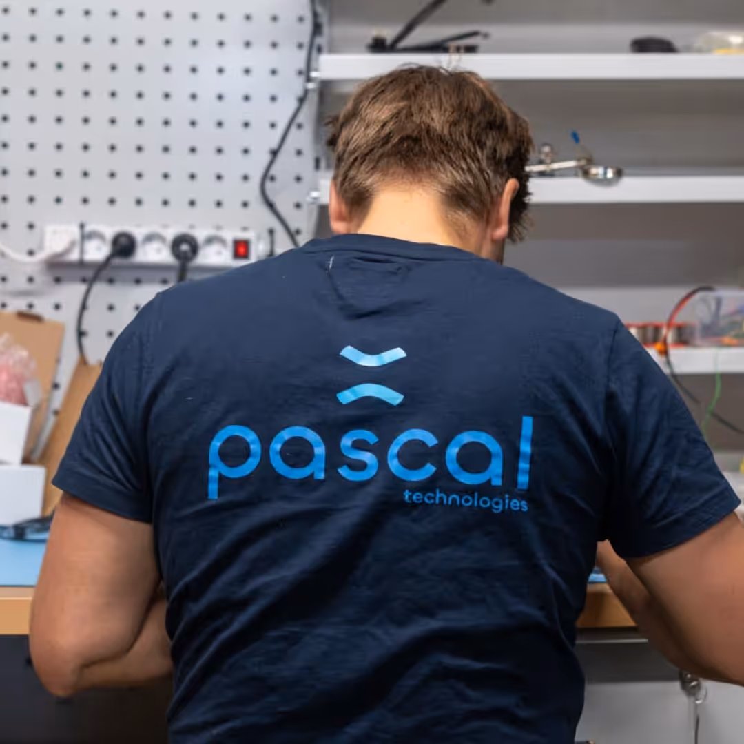 Person wearing a navy blue t-shirt with the text 'pascal technologies' on the back in a workshop.