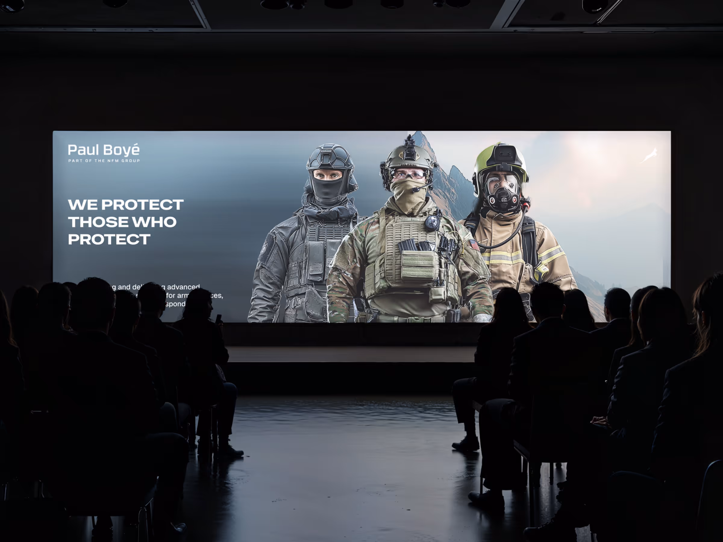 Konferansepublikum som ser på en stor skjerm med tre personer i militært og redningsutstyr og teksten 'We protect those who protect'.