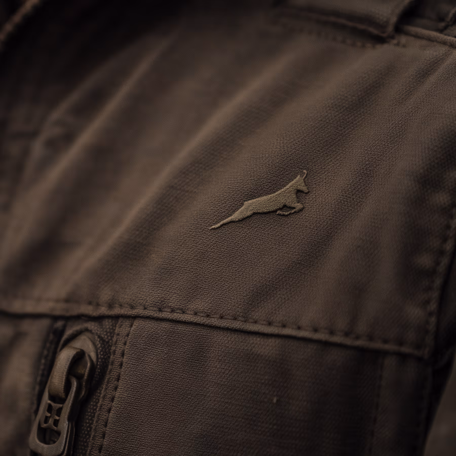 Detaljbilde av en mørk jakke med en brodert logo av et springende rovdyr over en glidelåslomme.