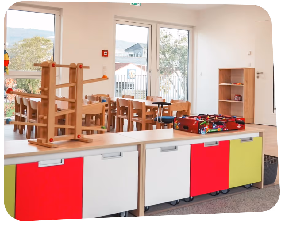 Bright classroom with wooden chairs and tables, colorful storage bins, a wooden toy, and large windows showing trees outside.