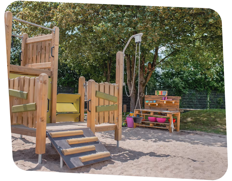 Children's playground with wooden climbing structure, swing, and sandpit, surrounded by trees and greenery.