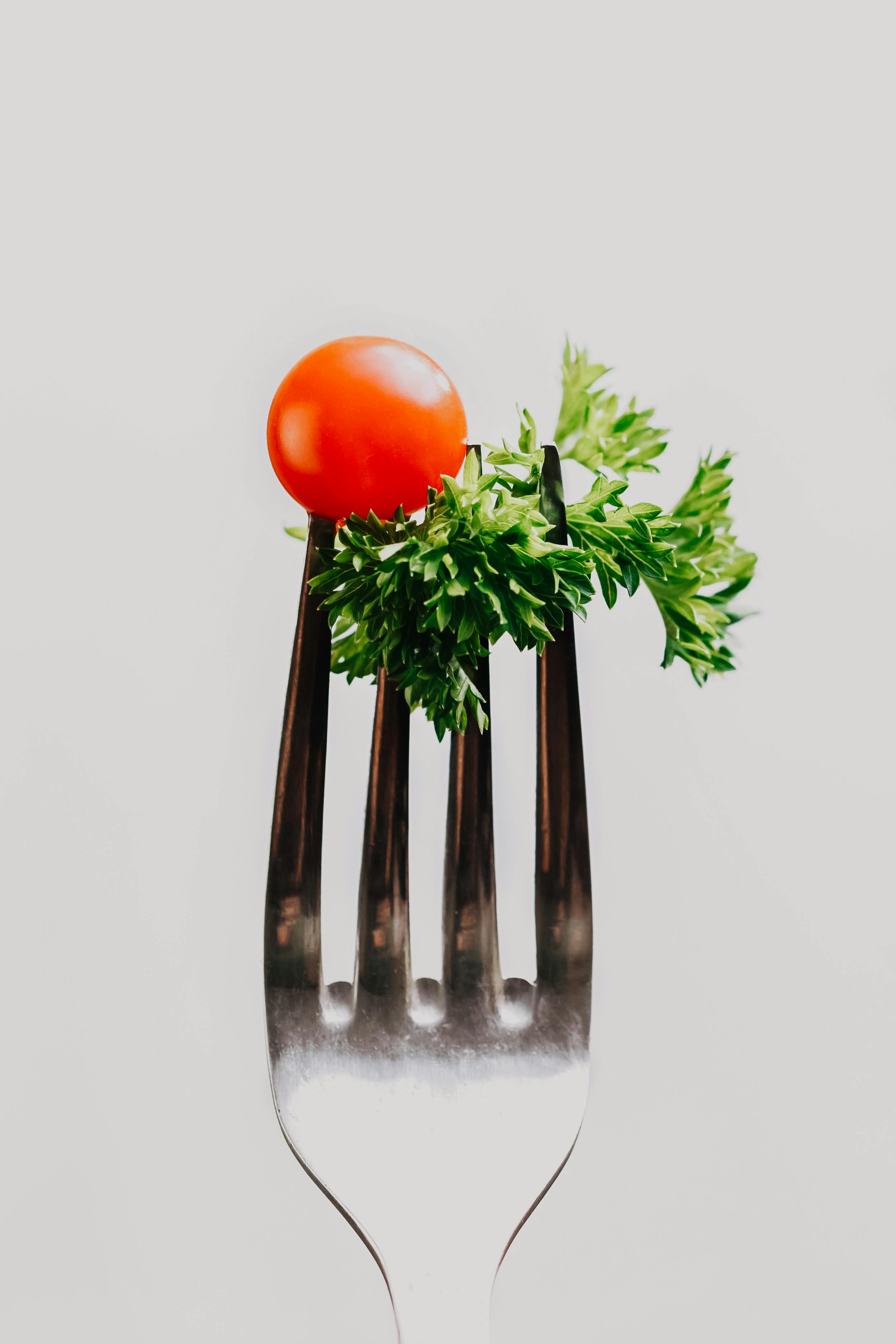 A silver fork holding a small red cherry tomato and green parsley leaves against a plain light background.