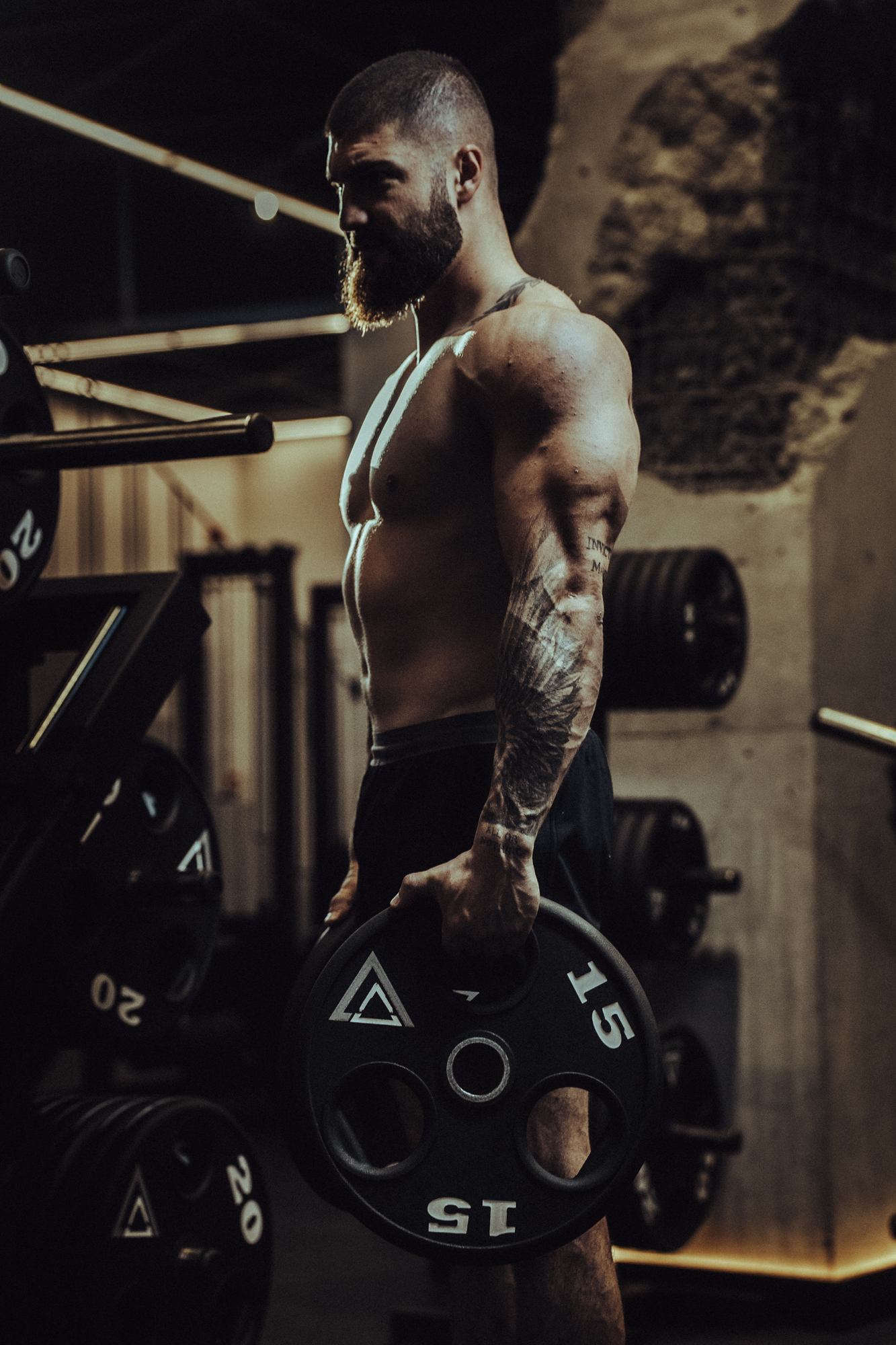 Muscular bearded man with tattoos lifting a 15-pound weight plate in a dimly lit gym.