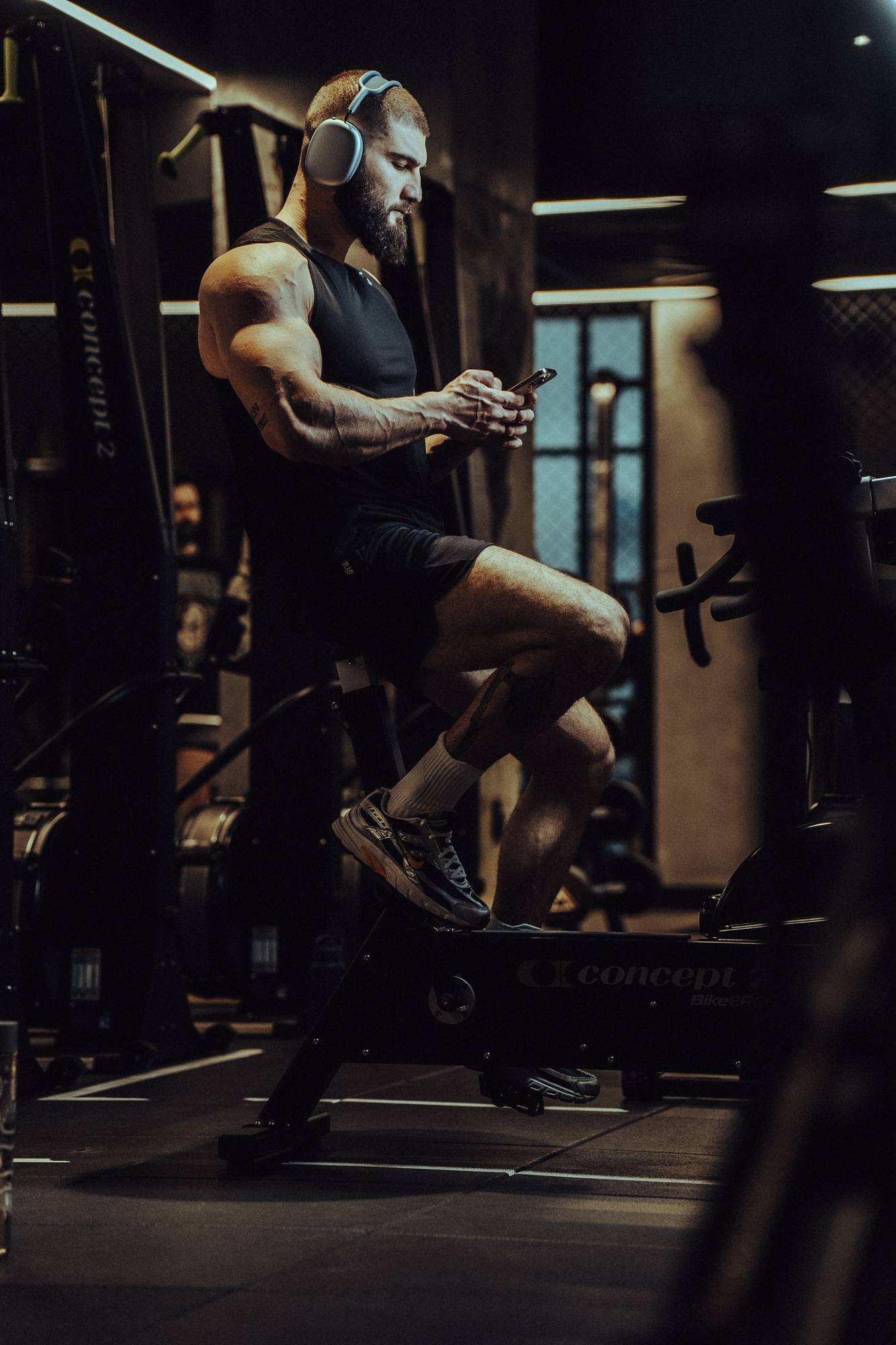 Muscular man wearing headphones sitting on a gym rowing machine while looking at his phone.