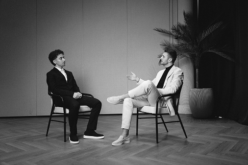 Twee mannen in pak zitten tegenover elkaar op stoelen in een kamer met houten vloer en een grote kamerplant.