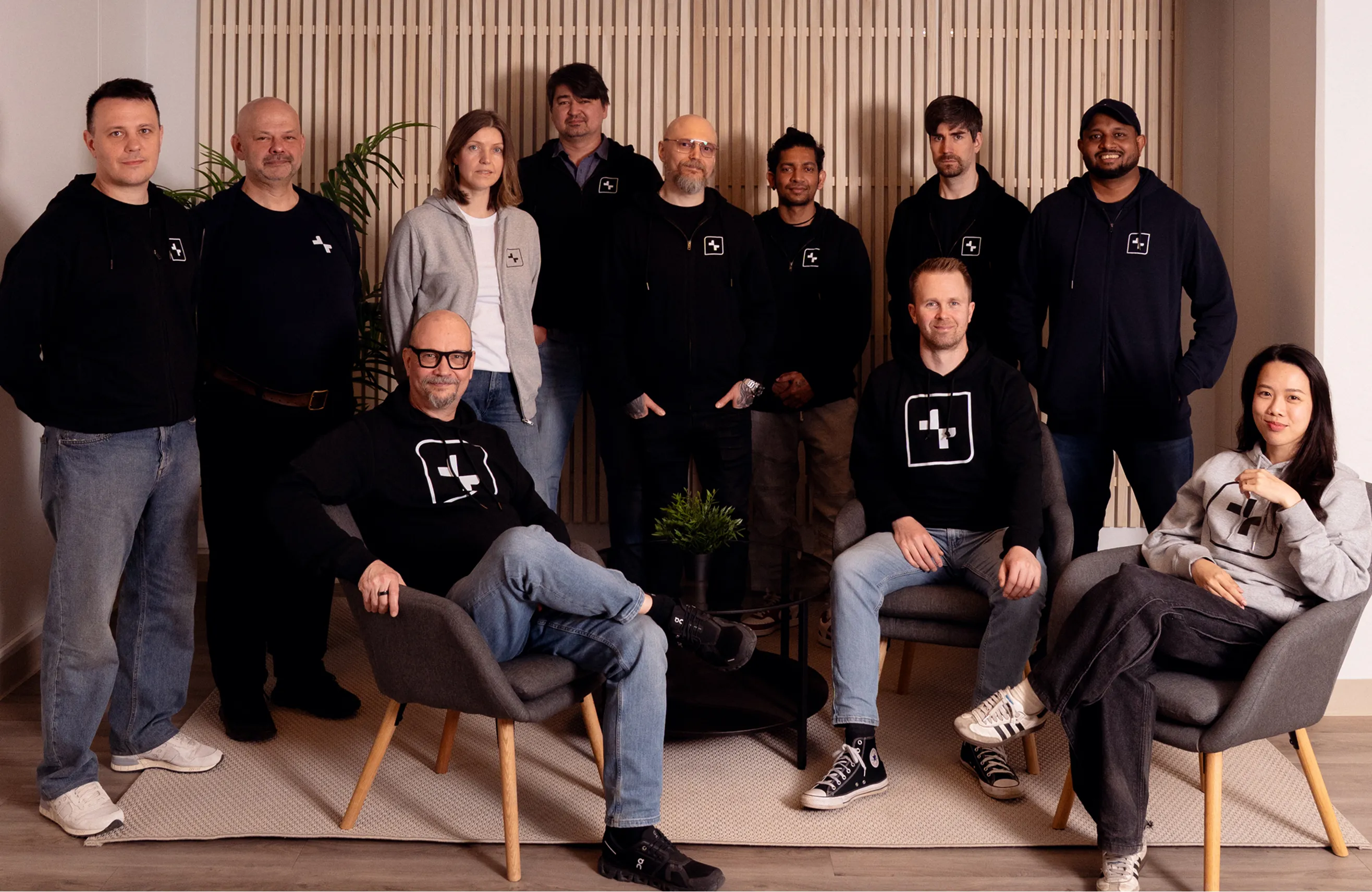 Group of eleven diverse adults posing indoors, most wearing black hoodies with logo, three seated in chairs.