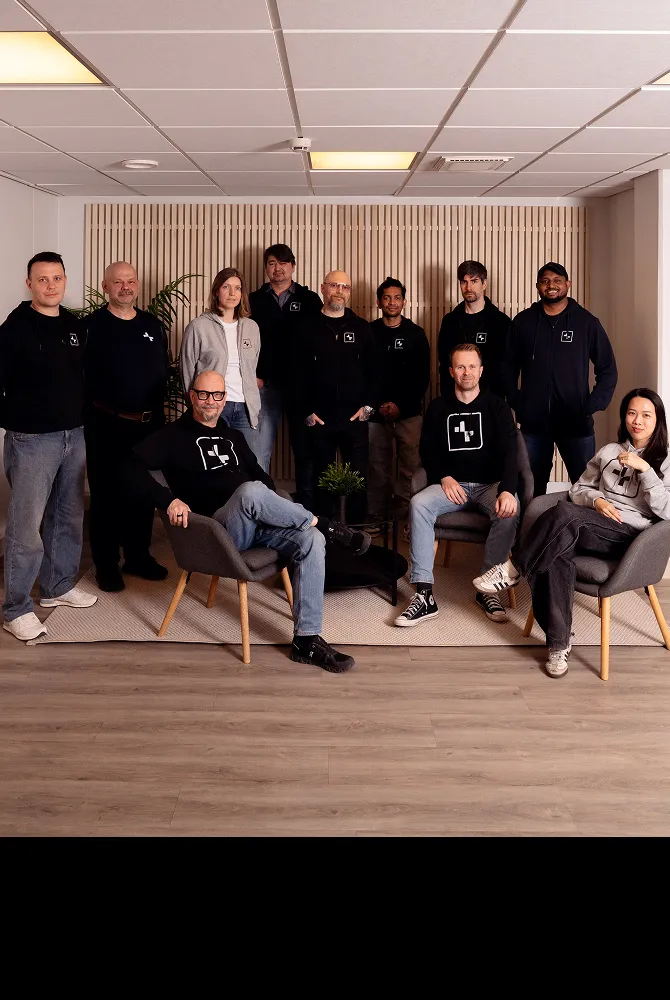 Group of eleven people posing in casual clothes in a modern office with wood panel background.