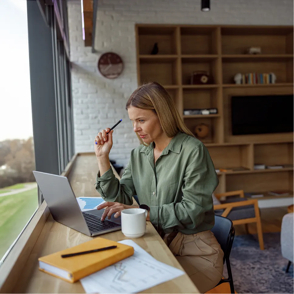 Woman with blonde hair wearing a green shirt working on a laptop at a wooden counter by a large window with papers, a yellow notebook, pen, and mug nearby.