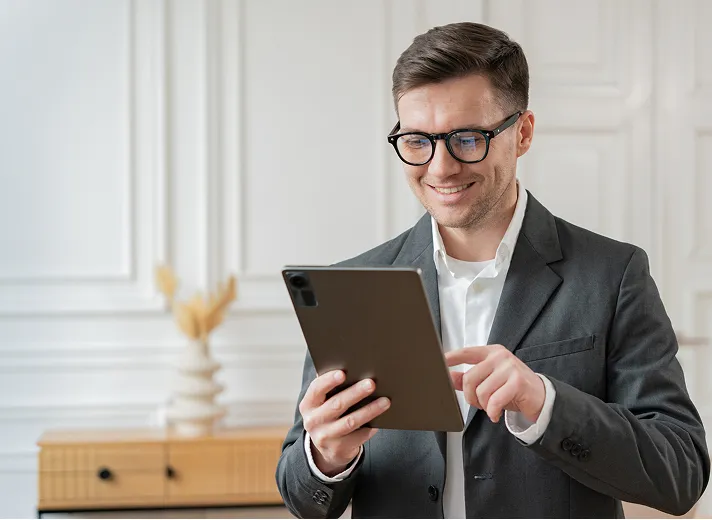 Smiling man in a dark suit and glasses using a tablet in a bright room with white walls and wooden furniture.