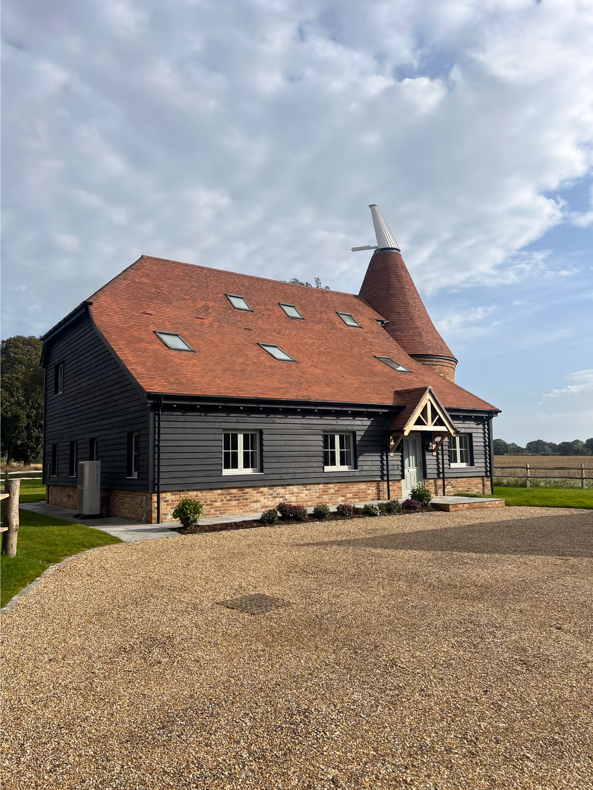 Modern house with black wooden siding, a red tiled roof with skylights, and a distinctive conical tower under a partly cloudy sky.