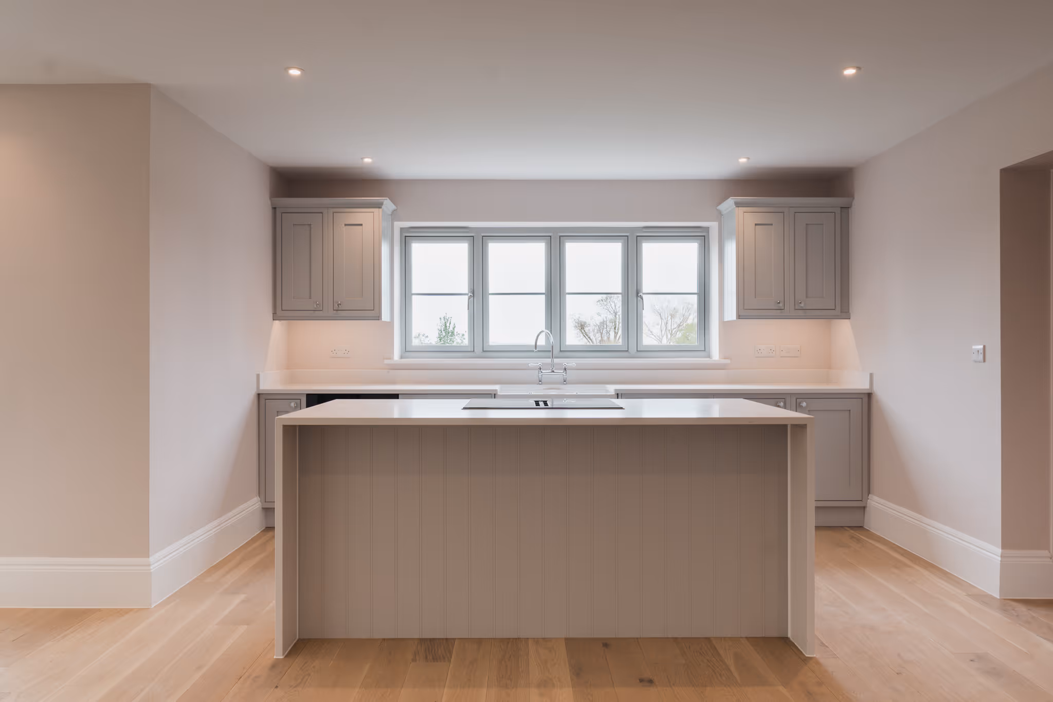 Minimalist modern kitchen with a large central island, light grey cabinetry, a sink under a four-panel window, and light wood flooring.