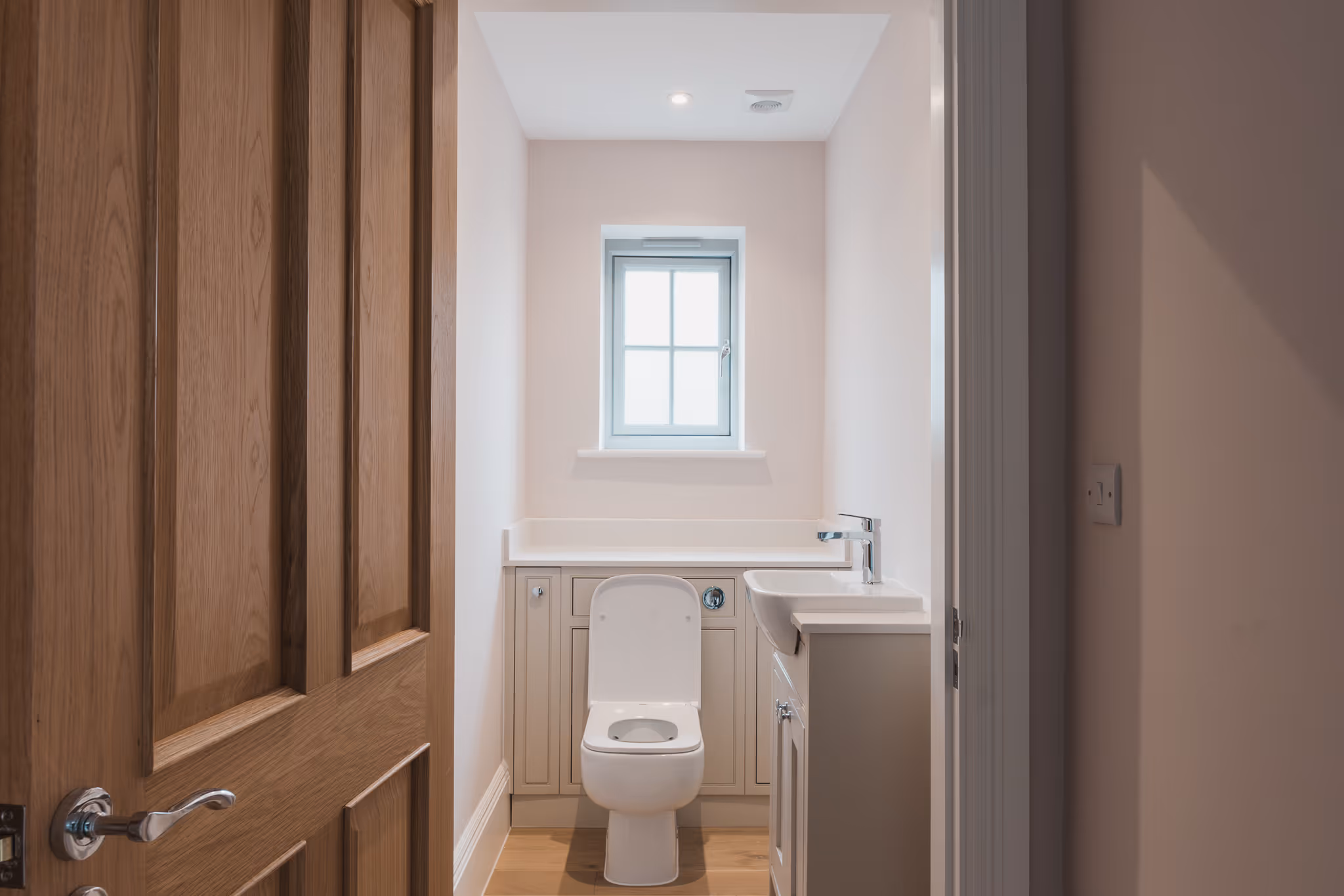 Small, modern bathroom with a white toilet, a sink with a silver faucet, light-colored walls, and a window above the toilet.
