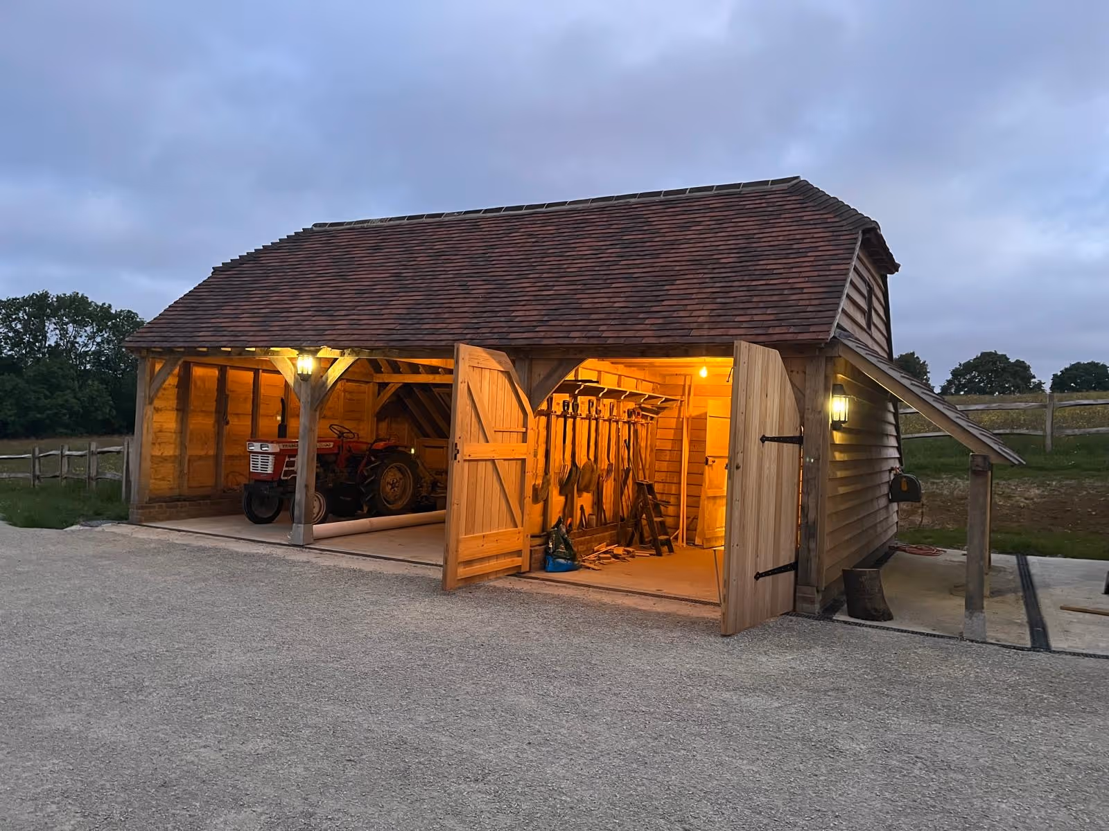 Wooden barn at dusk with open doors revealing a red tractor inside and farming tools hanging on the wall, illuminated by warm lights.