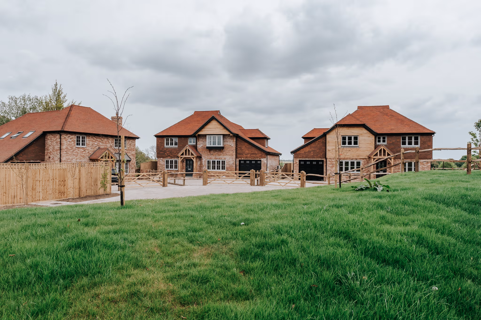 Three newly built two-story houses with brick and wood siding, red roofs, wooden fences, and a large green lawn in front under a cloudy sky.
