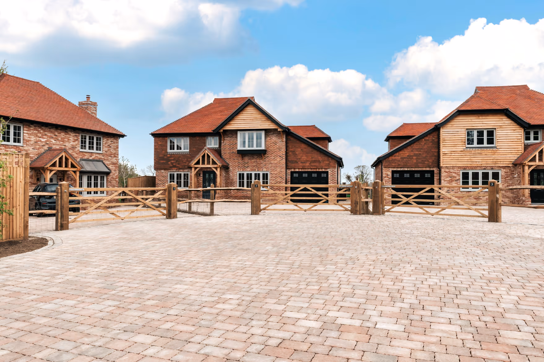 Three brick houses with red roofs, black garage doors, wooden gates, and paved driveway under a blue sky with clouds.