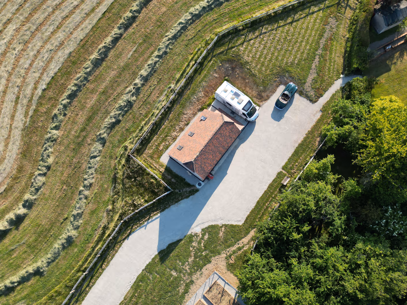 Aerial view of a small building with a red-tiled roof next to a parked camper van and a green convertible car, surrounded by fields and trees.