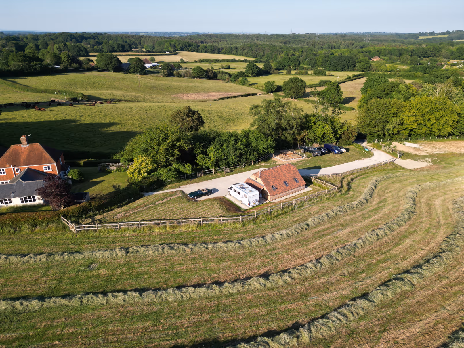 Aerial view of a rural landscape featuring a small brick house with a red roof, a white camper van, several parked cars, fenced fields, and green pastures extending into the distance under a clear sky.