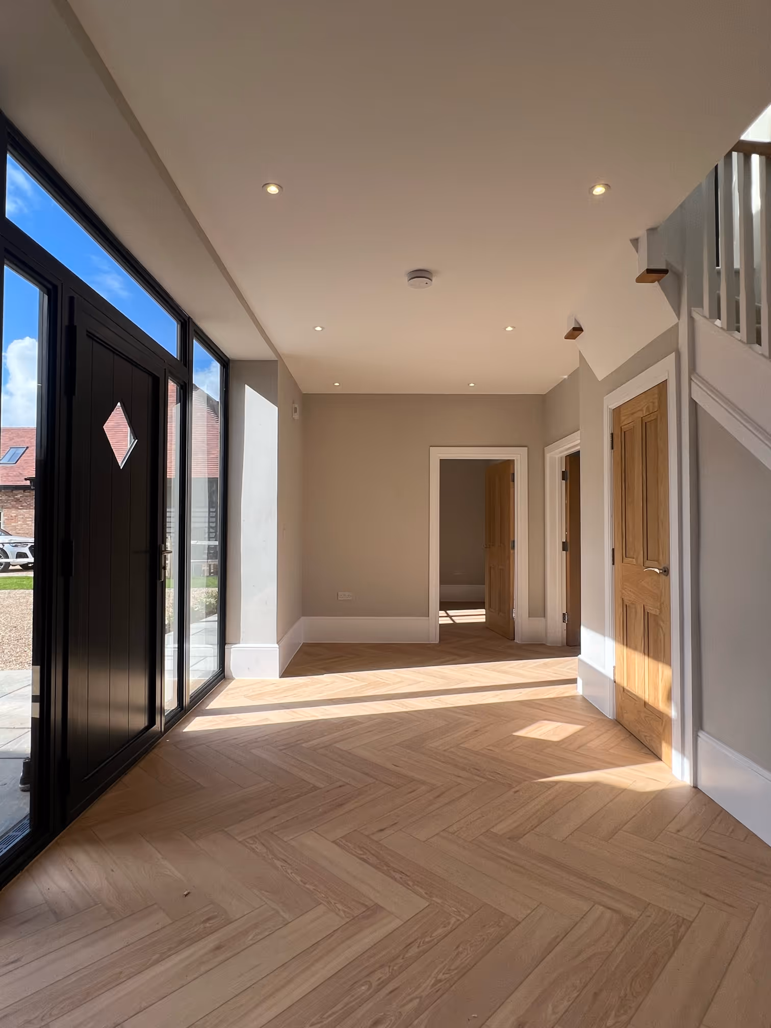 Bright empty room with light wood herringbone floor, beige walls, recessed ceiling lights, black glass door on the left, and three wooden doors on the right under a staircase.
