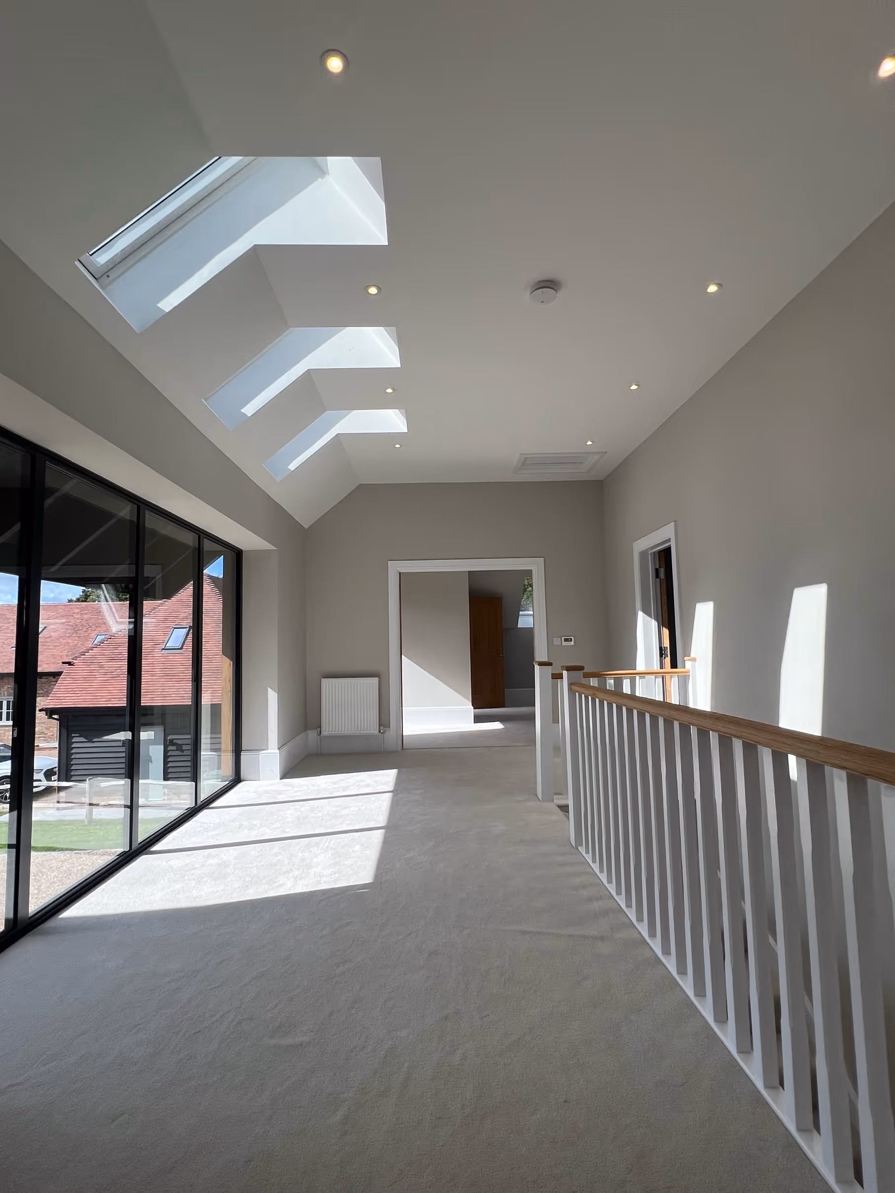 Bright hallway with large skylights, recessed ceiling lights, glass sliding doors on the left, and a white wooden railing on the right.