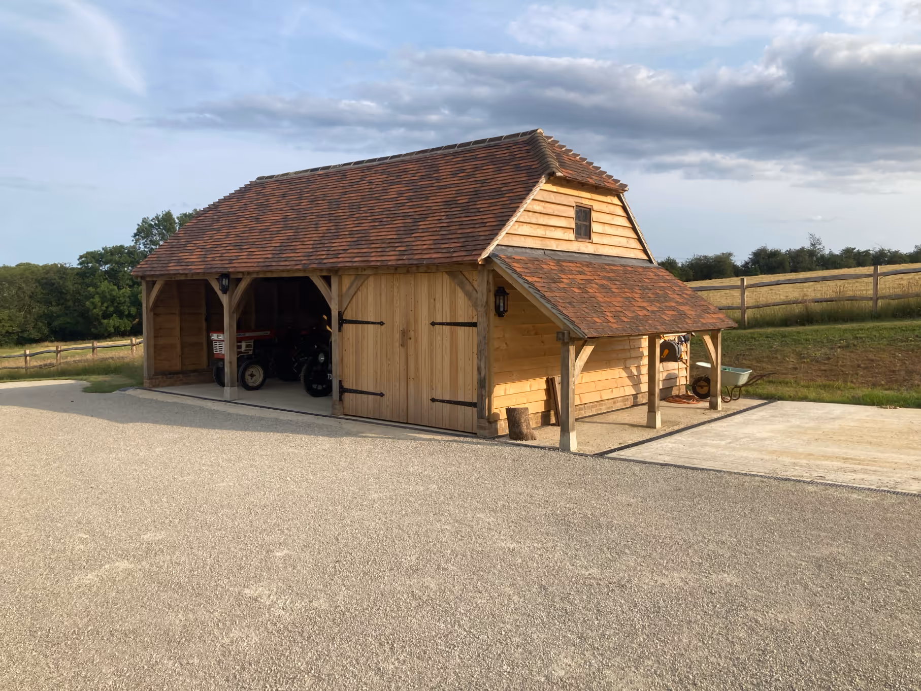 Wooden barn with a tiled roof housing tractors, set on a gravel area with fields and a wooden fence in the background.