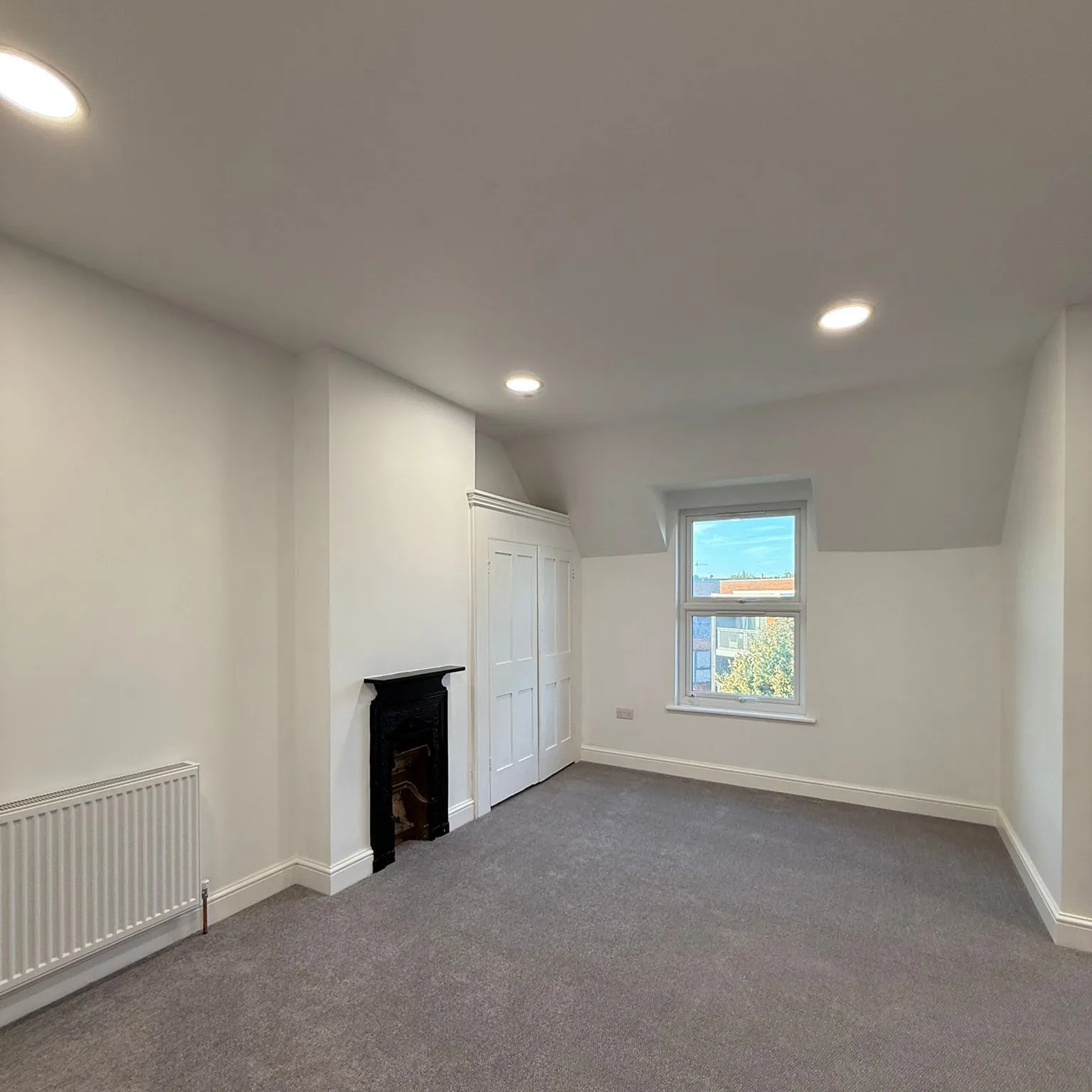 Empty room with white walls, gray carpet, recessed ceiling lights, a small fireplace, built-in white closet, and a single window showing an outdoor view.