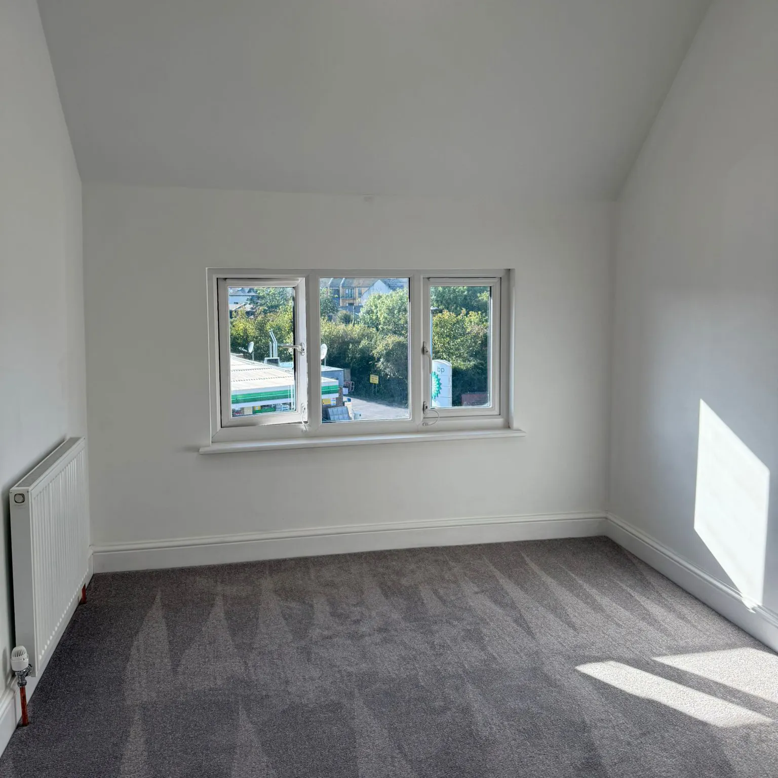 Empty room with gray carpet, white walls, a radiator on the left, and three-pane window overlooking trees and a gas station.