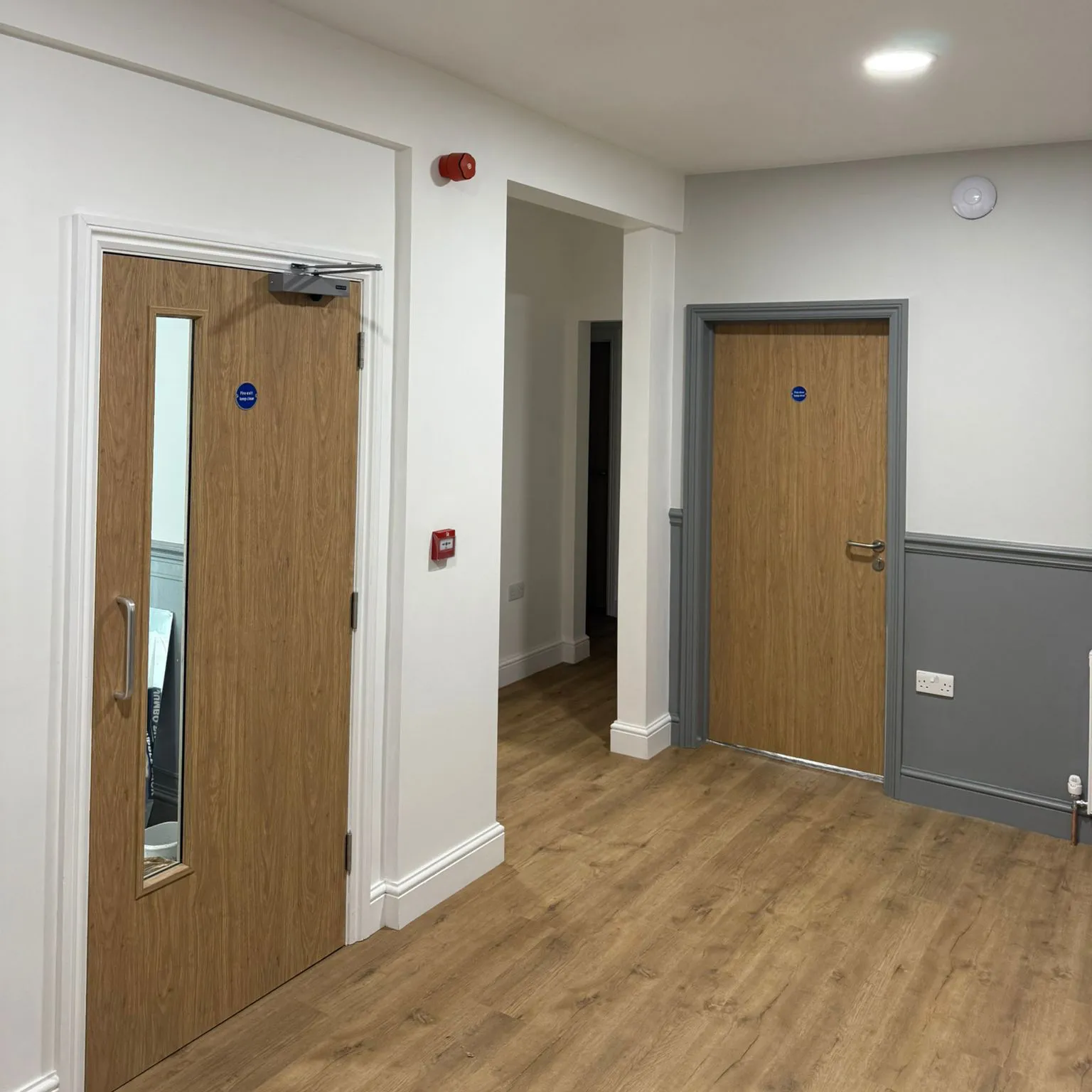 Interior hallway with two closed wooden doors, one with a vertical window and white walls with gray wainscoting and wood-look flooring.