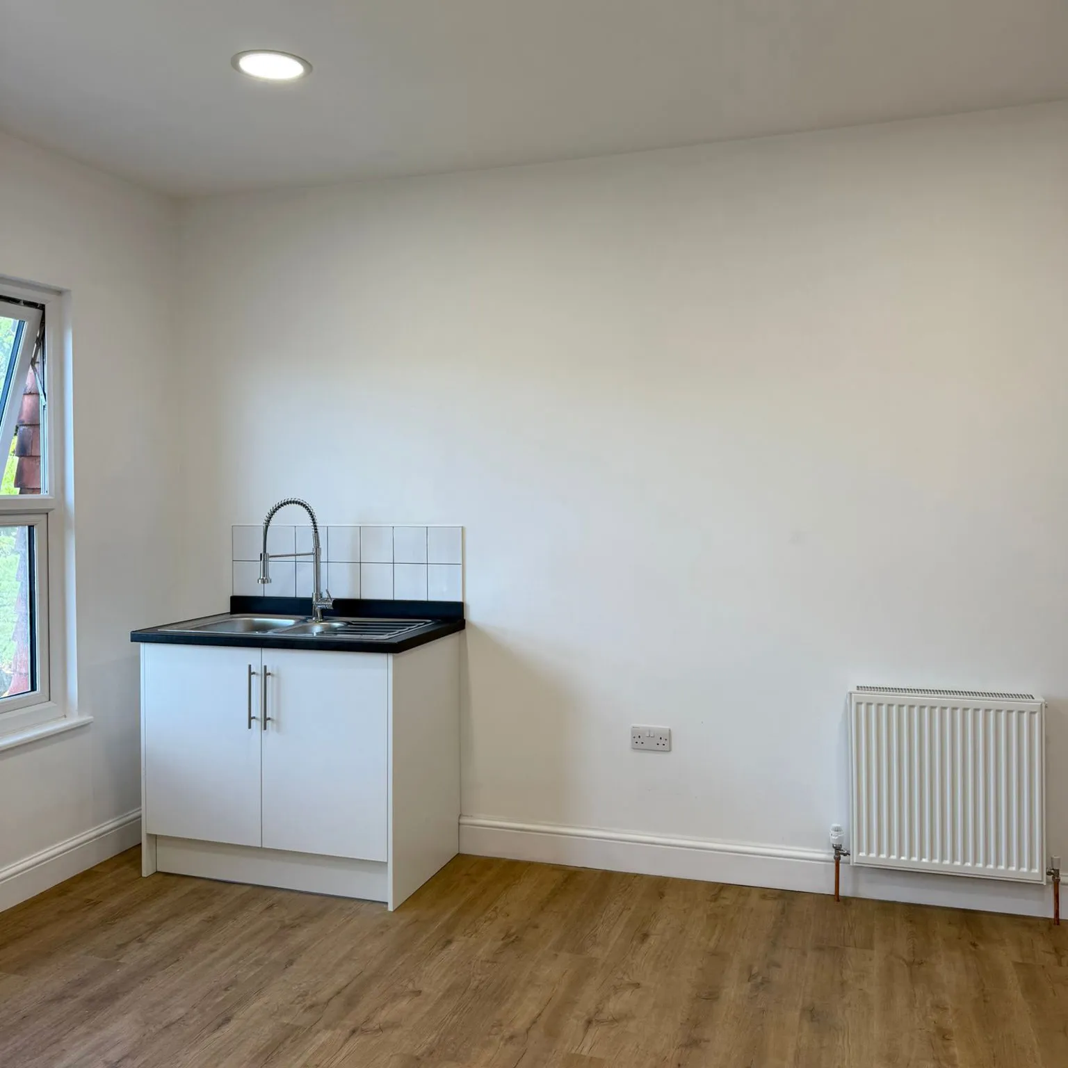 Minimalist kitchen corner with white cabinet, black countertop, stainless steel sink, window, and radiator on wooden floor.
