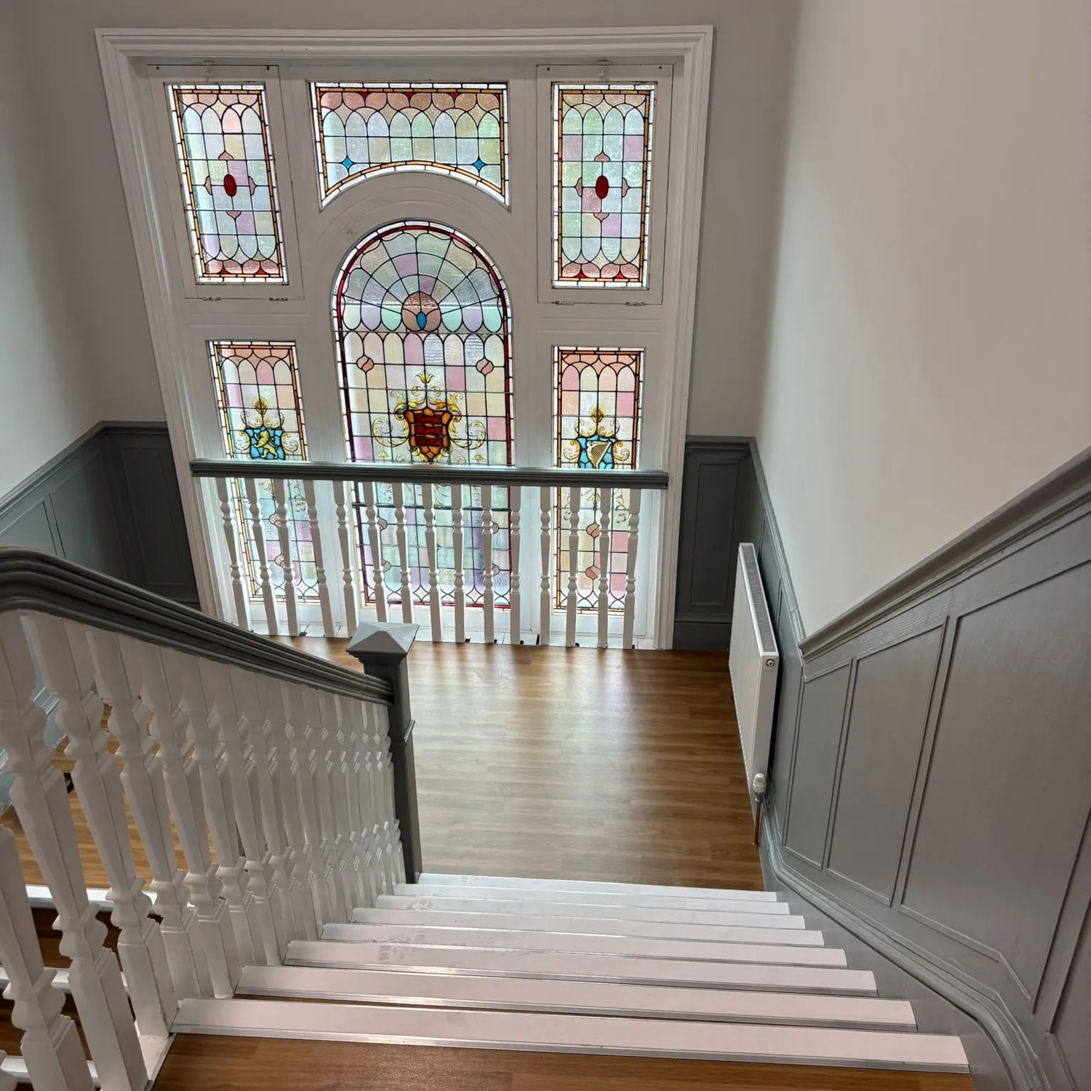 Top view of a staircase with white railing and wooden steps leading down to a landing that features a large decorative stained glass window with colorful patterns and crests.