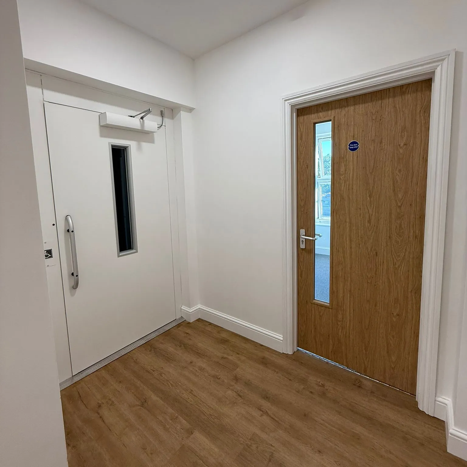 Interior corner with a white accessibility door and a wooden door with a vertical window showing a room with windows.