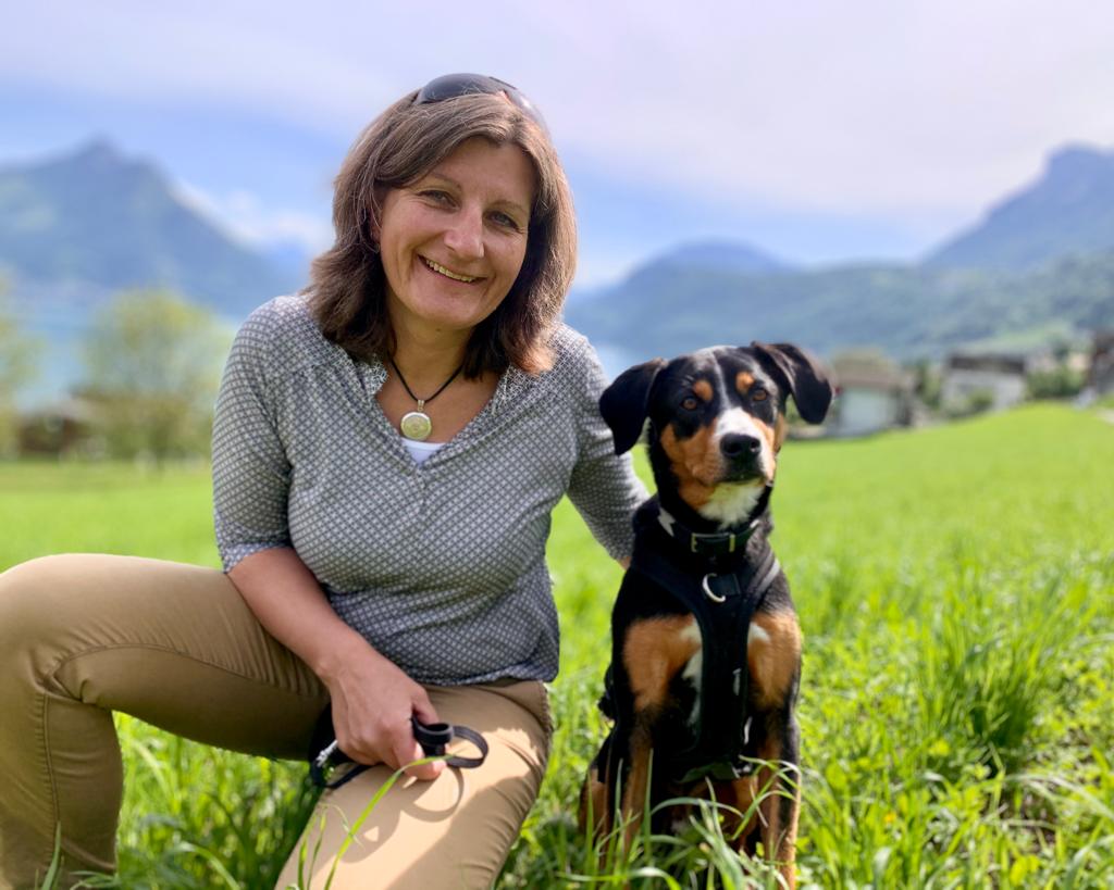 Lächelnde Ursula Hess  kniet auf Wiese neben schwarzem Hund mit braunen Abzeichen vor Berglandschaft.