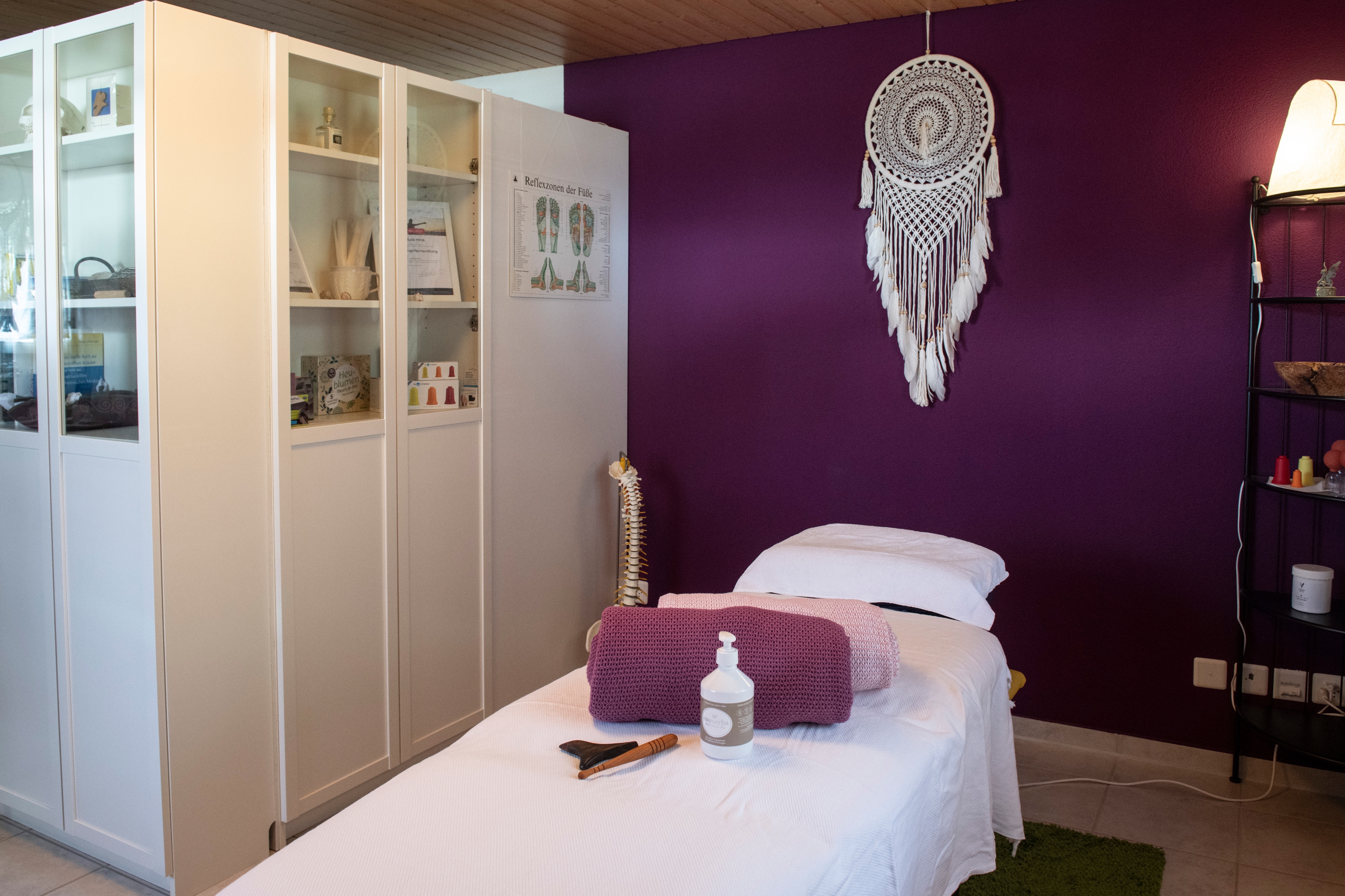 Behandlungsraum mit Massagetisch, violetter Wand, Traumfänger, weissen Schränken und Regalen mit Massagezubehör.
