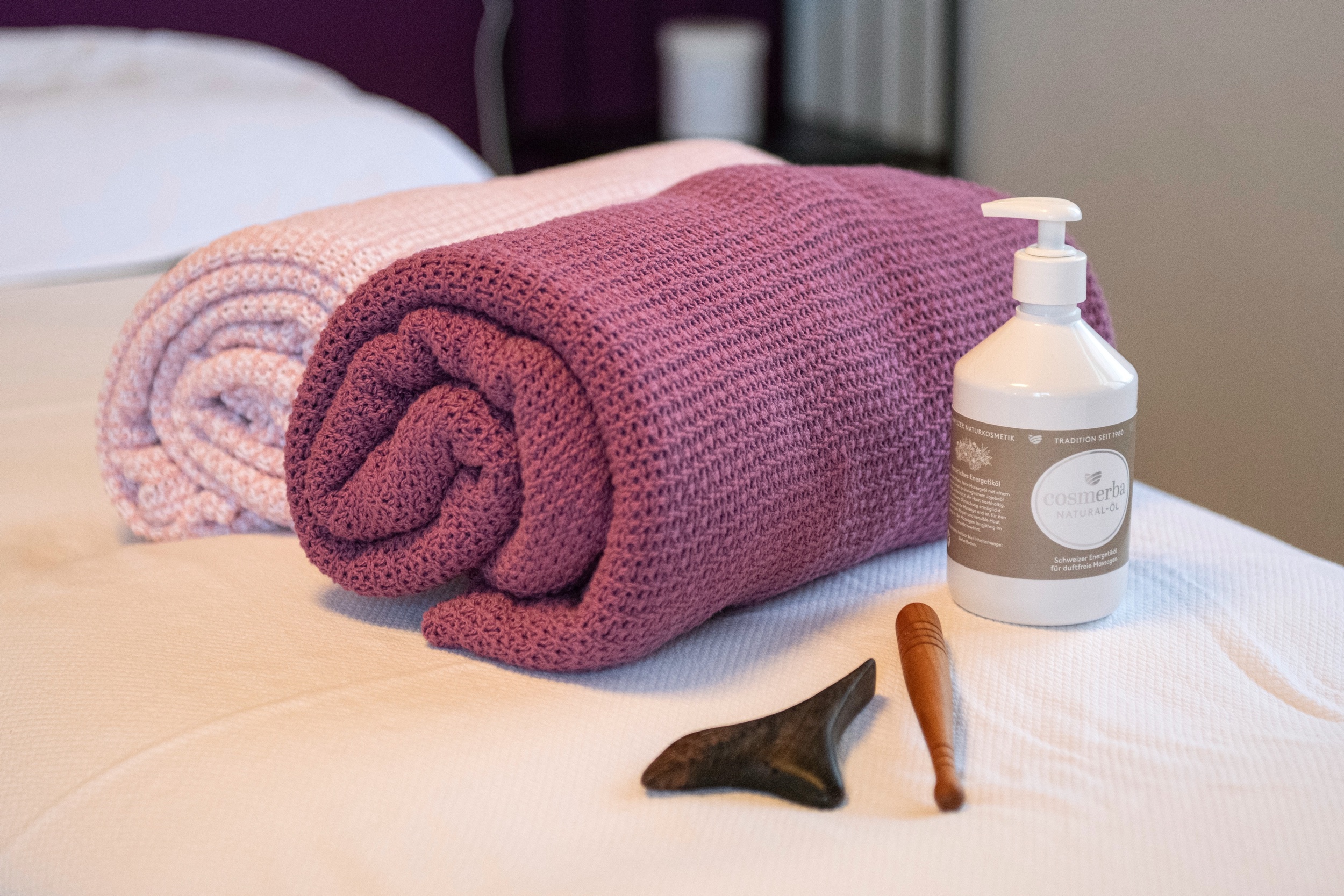 Zwei eingerollte gestrickte Handtücher auf einem weißen Bett, daneben eine weiße Flasche Cosmerba Natural-Öl und zwei Massagewerkzeuge.